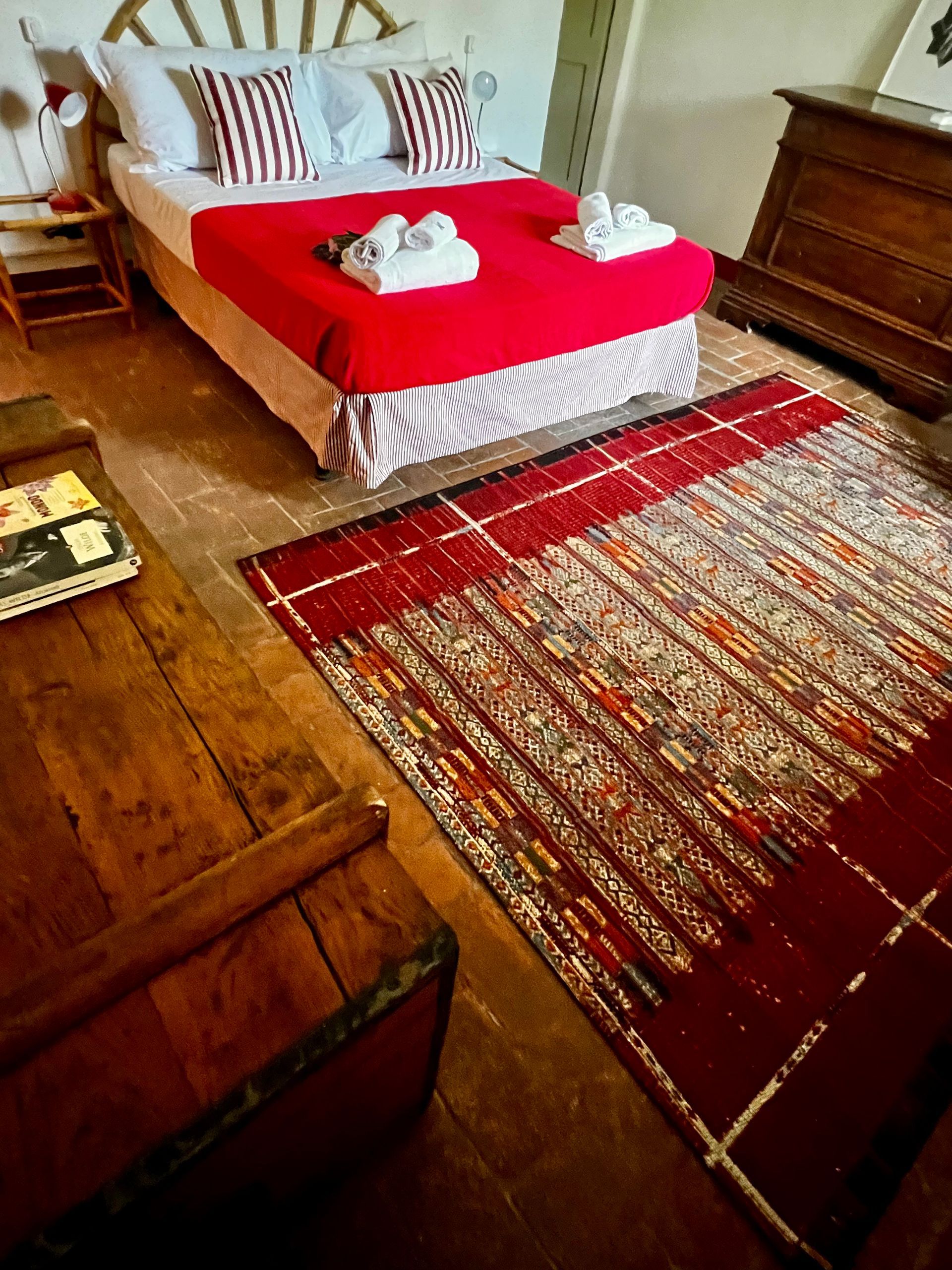 Una camera da letto presenta un letto con lenzuola rosse, un tappeto a motivi geometrici, una cassapanca di legno in primo piano e una cassettiera di legno.