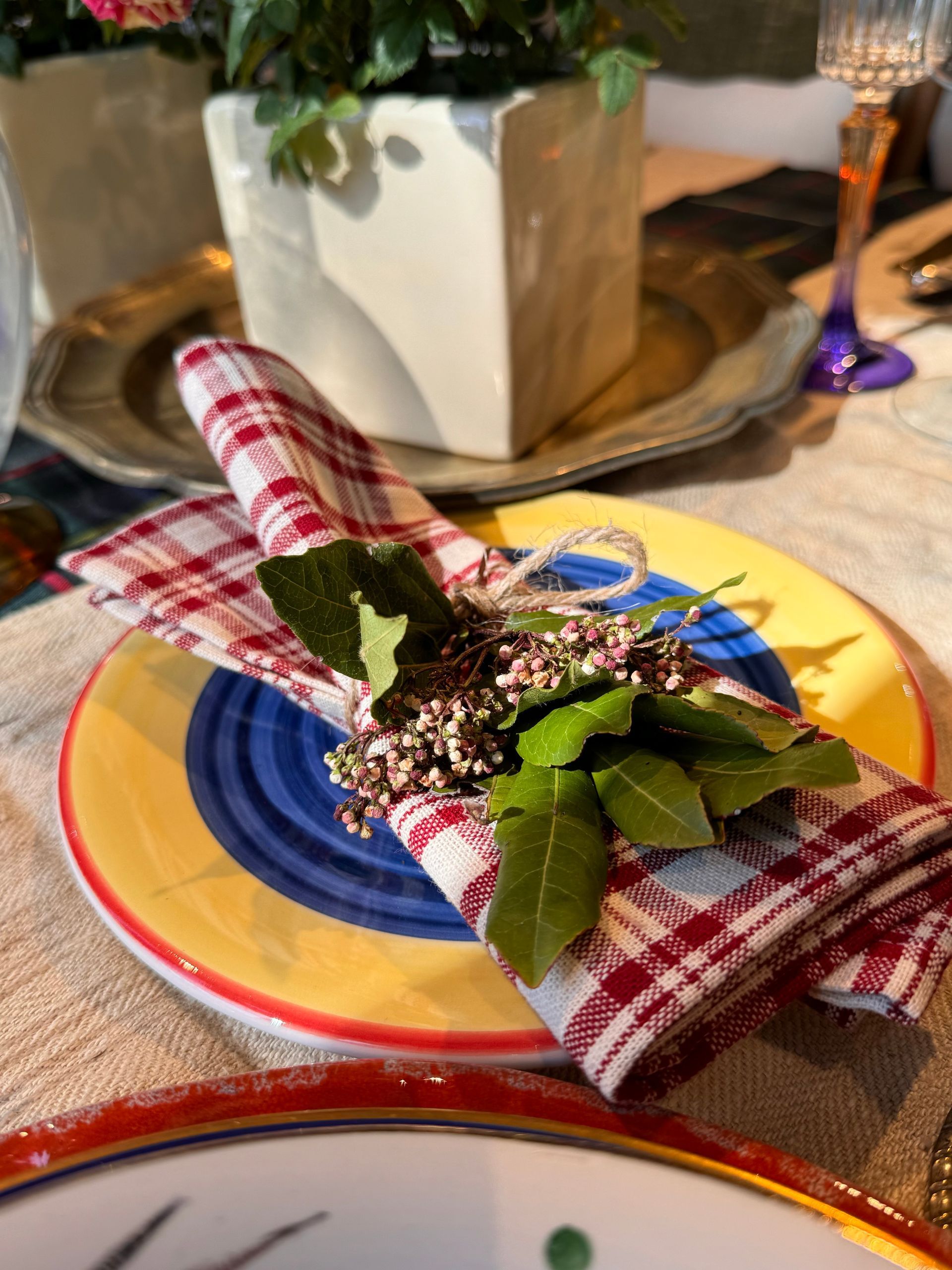 Una mise en place con un tovagliolo a quadri rossi e bianchi legato con verde e piccoli fiori bianchi su un piatto colorato.