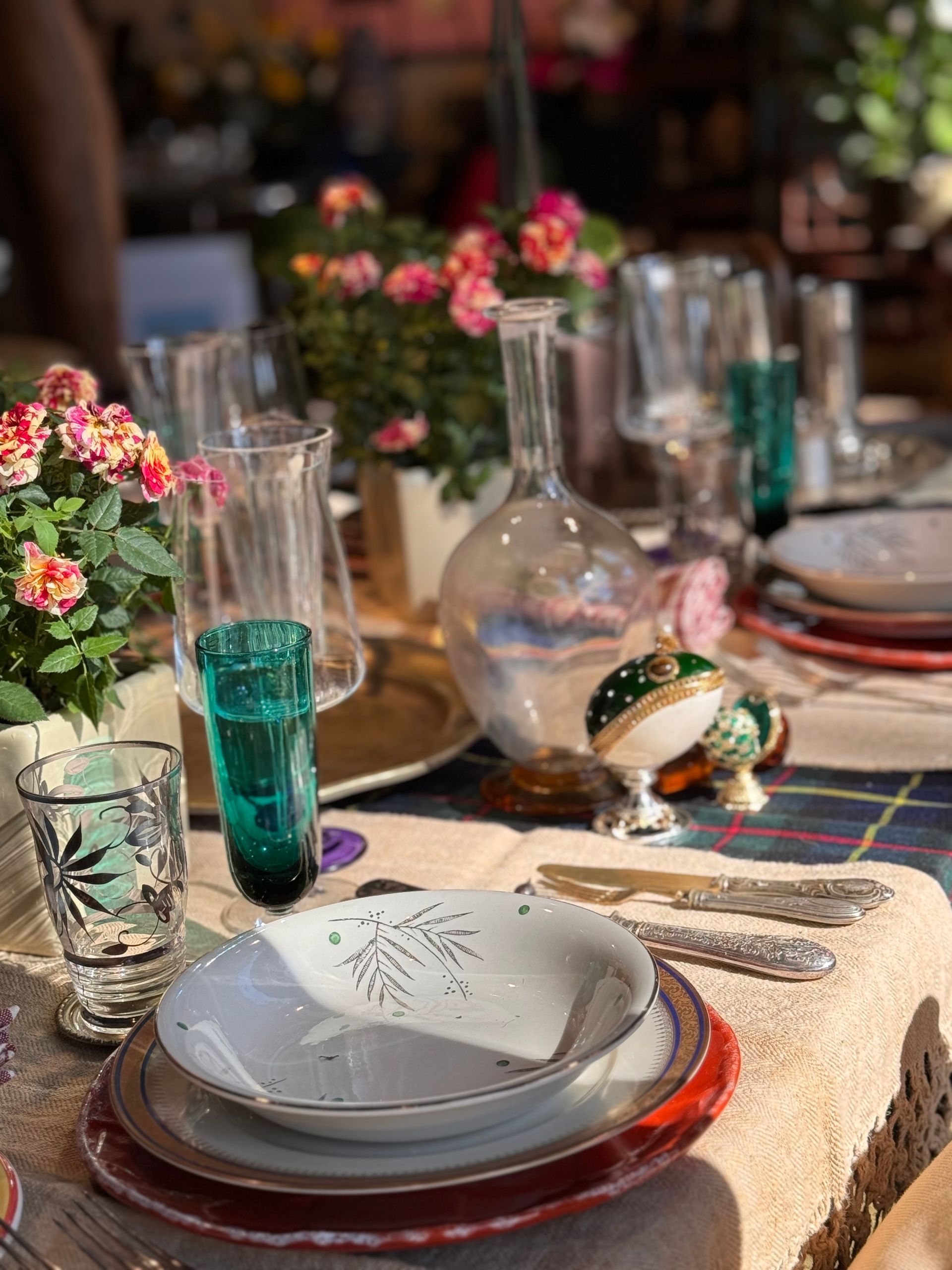Una tavola apparecchiata con posate, vasi decorativi, fiori freschi e bicchieri vintage su una tovaglia a quadri.