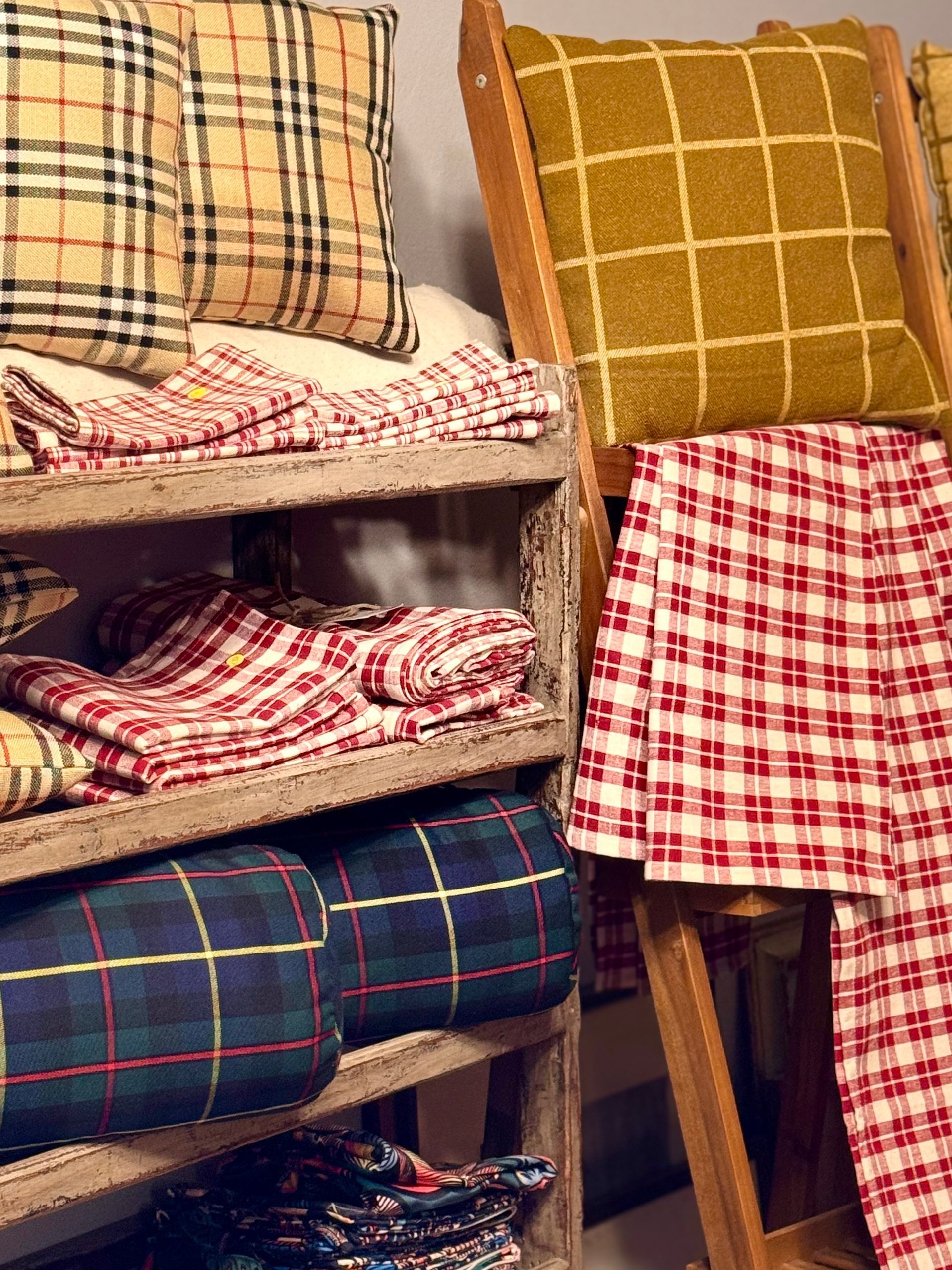 Una scaffalatura in legno espone diverse coperte e cuscini a quadri e scacchi nei toni del beige, del rosso e del blu.