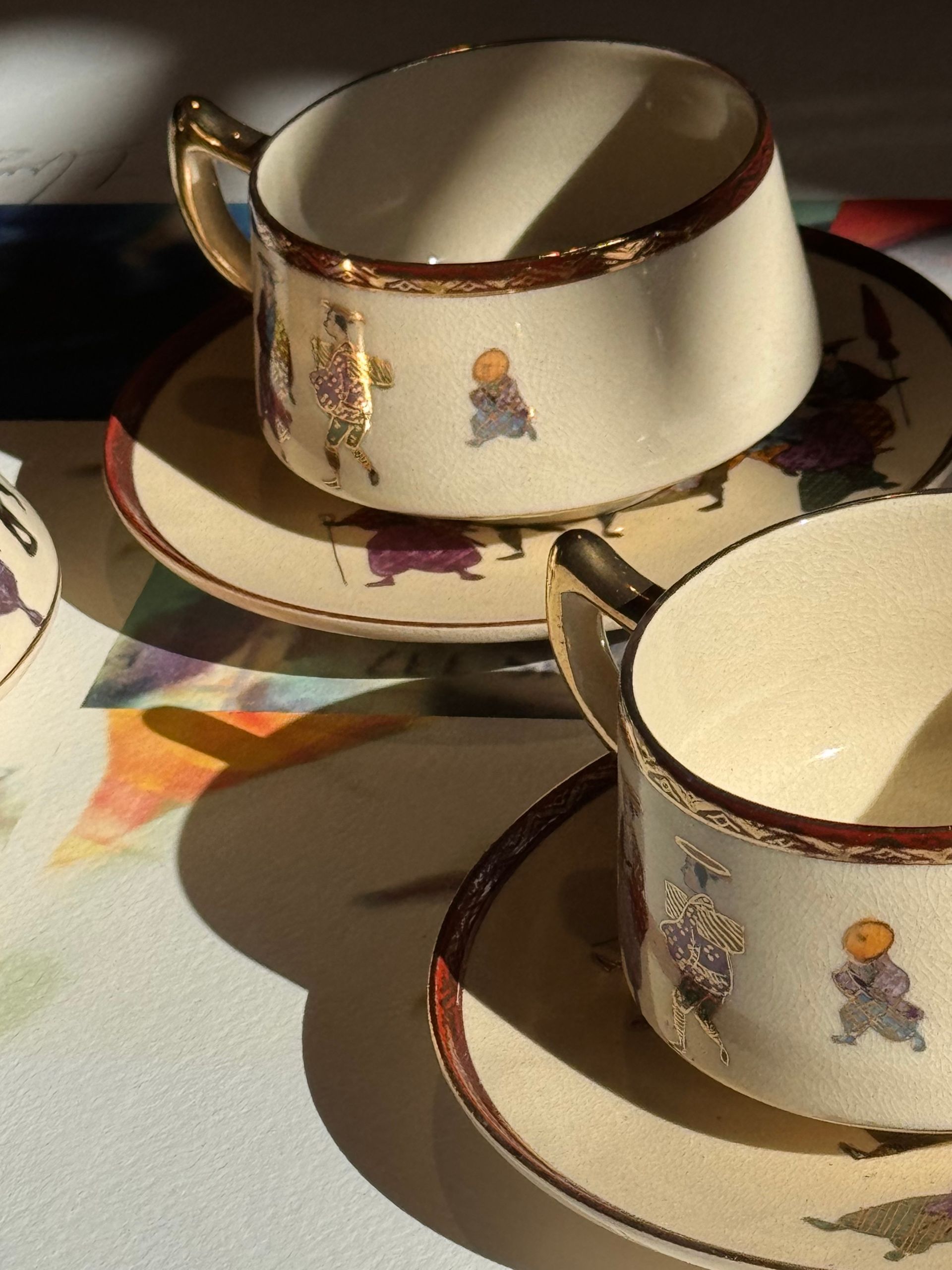 Due tazze da tè color crema con piattini abbinati, decorate con piccole figure e bordi dorati, sono esposte alla luce del sole.