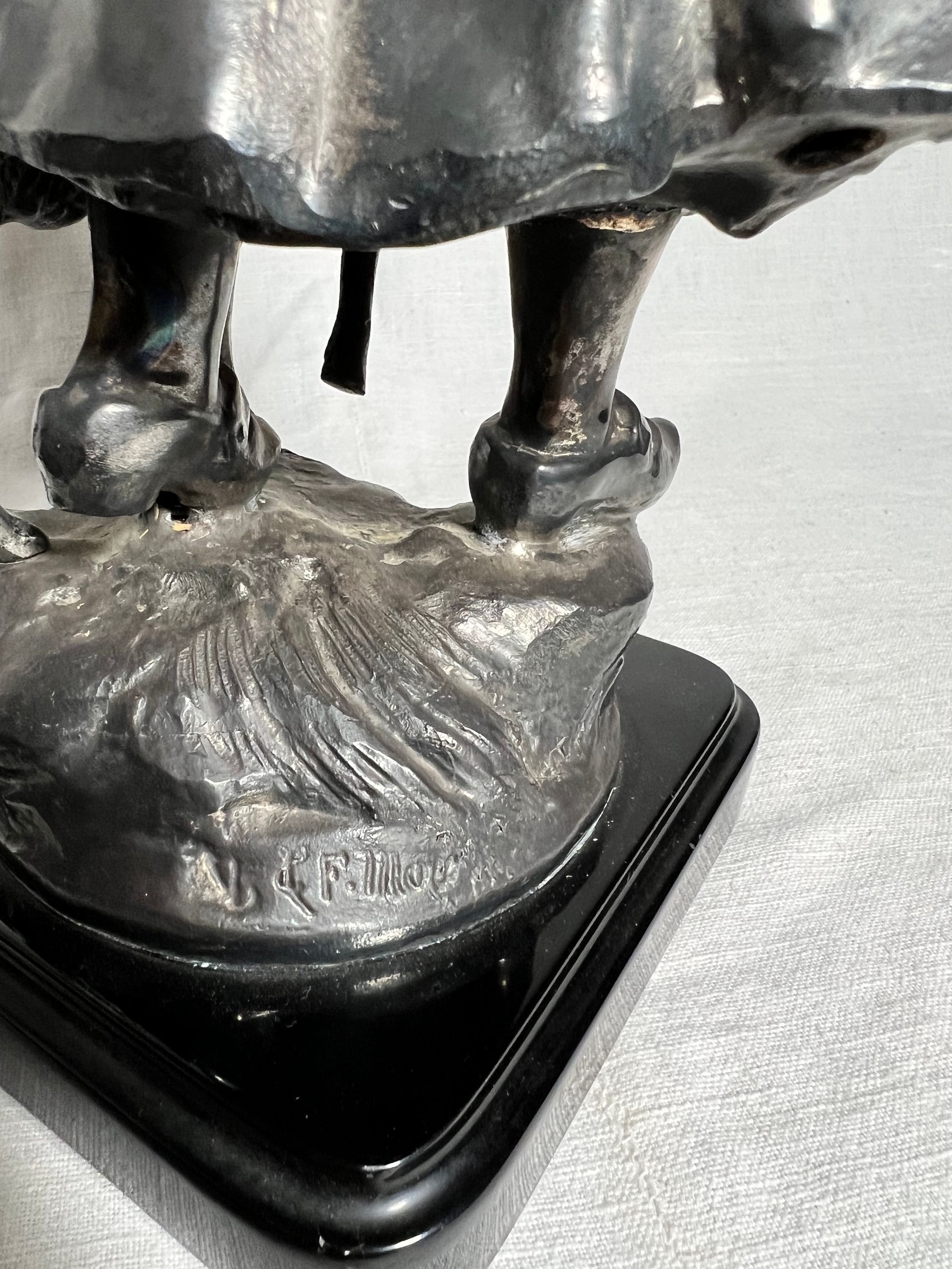 Primo piano della base di una scultura metallica con dettagli modellati e firma incisa su un piedistallo nero.