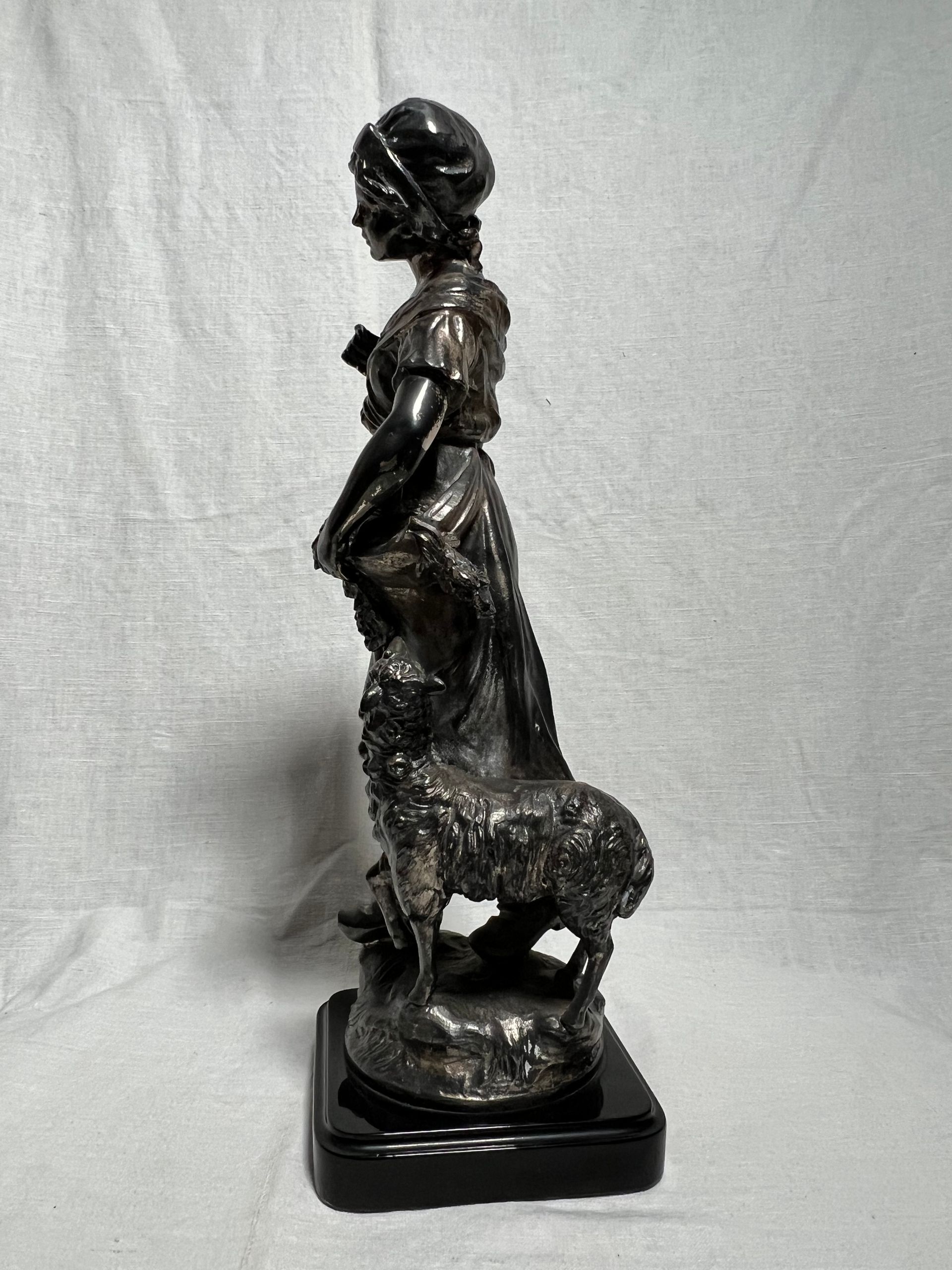 Una statua scura e metallica raffigurante una persona in piedi accanto a una pecora, montata su una base quadrata nera su uno sfondo drappeggiato.