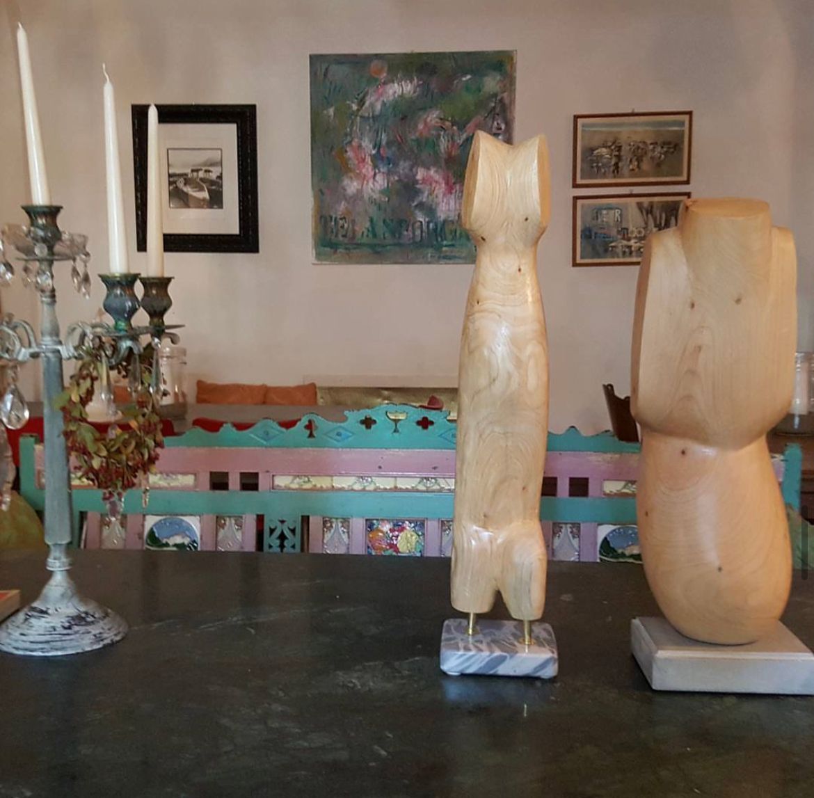 Due sculture in legno sono posizionate su un tavolo accanto a un portacandele, di fronte a una panchina colorata e a un'opera d'arte appesa al muro.