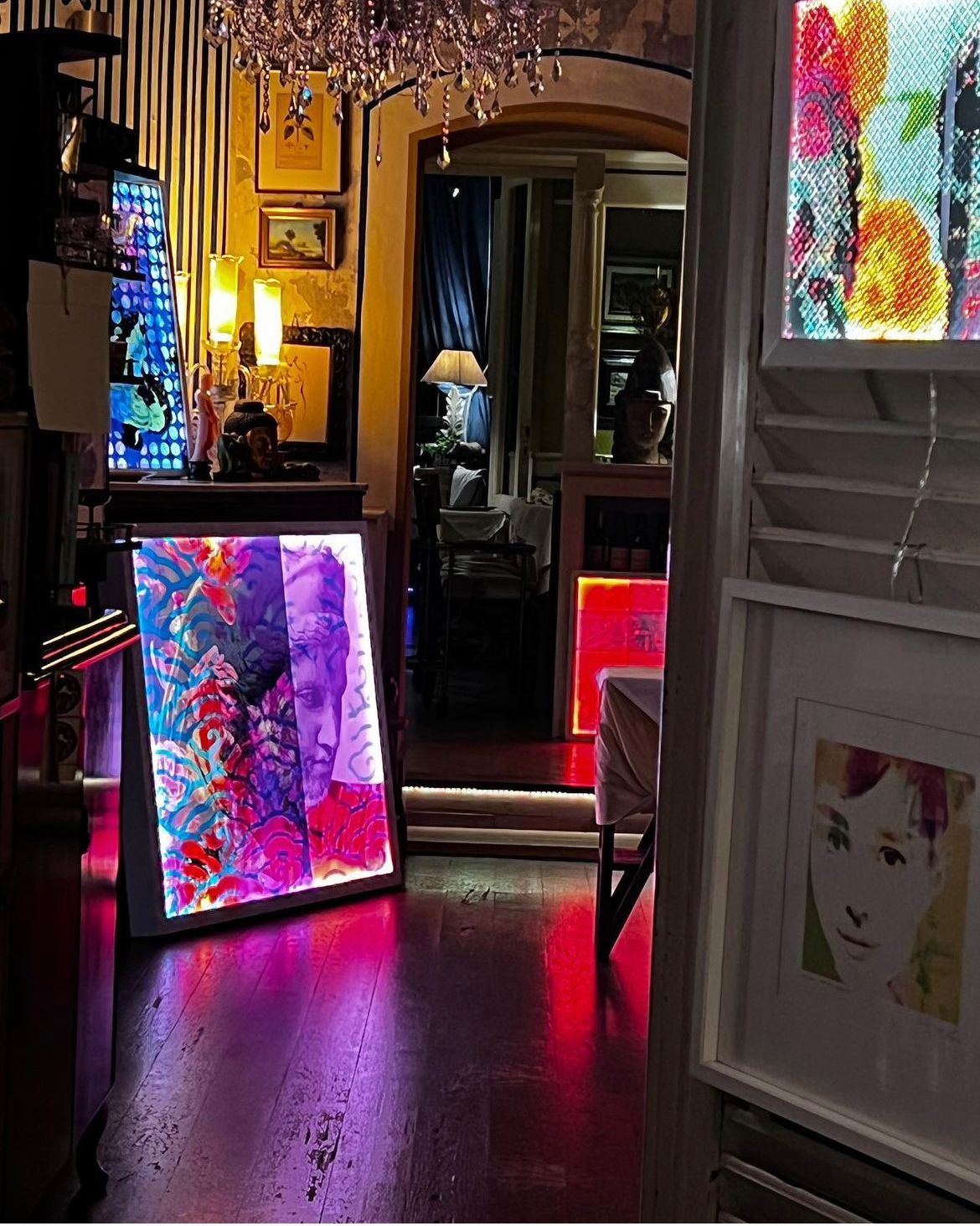 Una stanza illuminata da una luce calda, con un'opera d'arte vibrante e luminosa sul pavimento e un ritratto incorniciato di un volto sulla parete di destra.