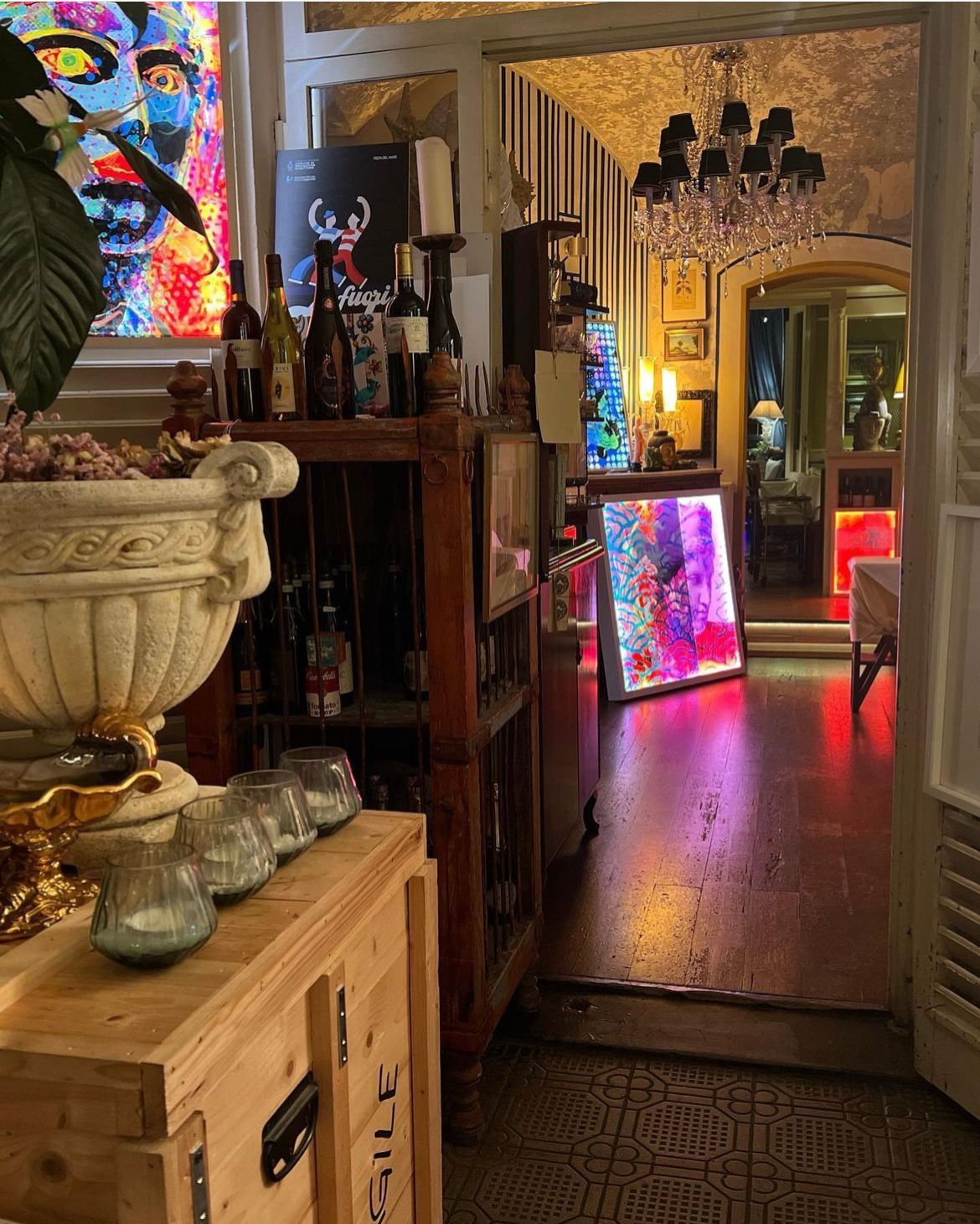 Una stanza eclettica e in penombra, con vivaci opere d'arte luminose alle pareti, bottiglie di vino su un supporto di legno e un'illuminazione calda e colorata.