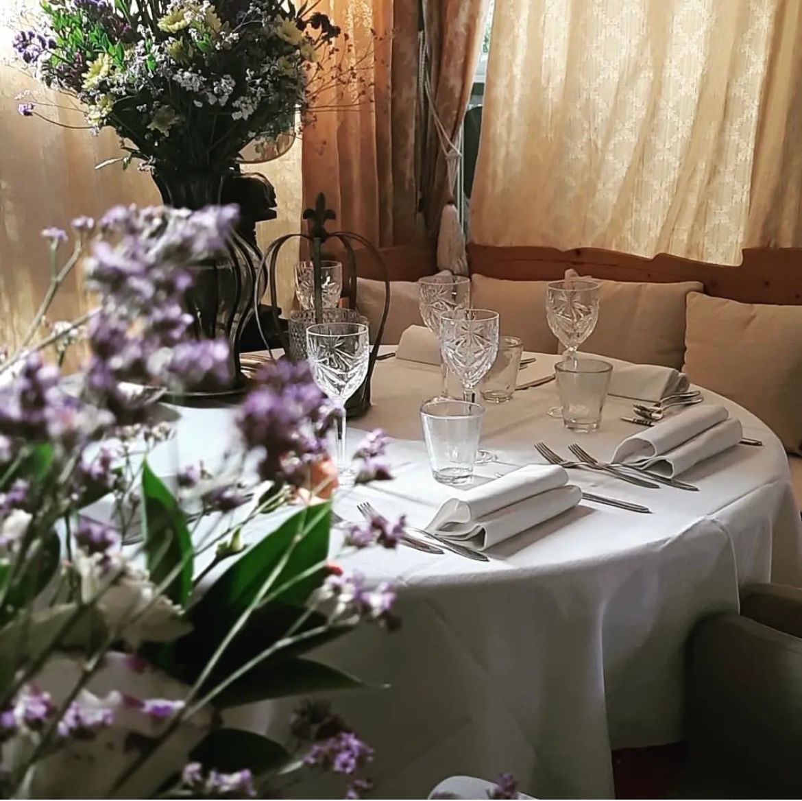 Un tavolo da pranzo rotondo apparecchiato con tovaglie bianche, bicchieri di cristallo e posate d'argento, con fiori sfocati in primo piano.