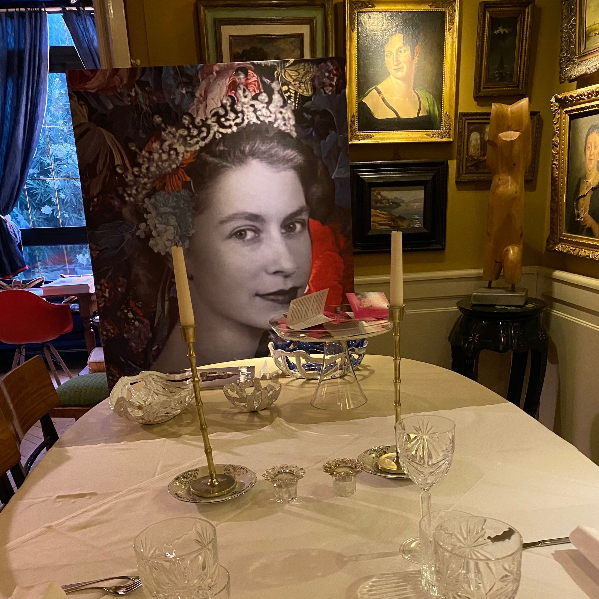 Su un tavolo da pranzo apparecchiato con candele, bicchieri e tovaglie bianche, è appoggiato un ritratto incorniciato della regina Elisabetta II con indosso una corona.