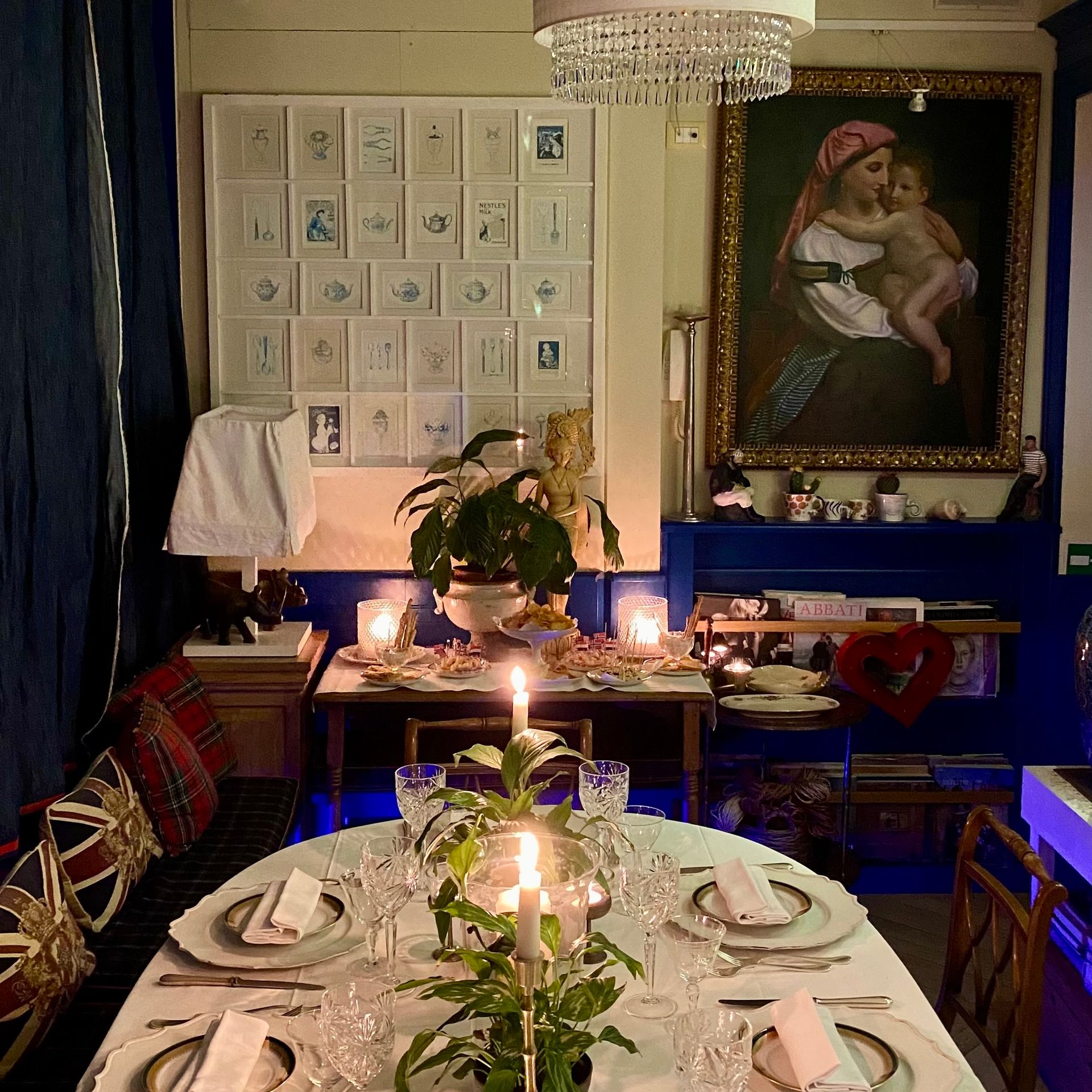 Una sala da pranzo apparecchiata per la cena con candele accese, fiori, porcellane pregiate e un grande dipinto a olio su una parete blu scuro.