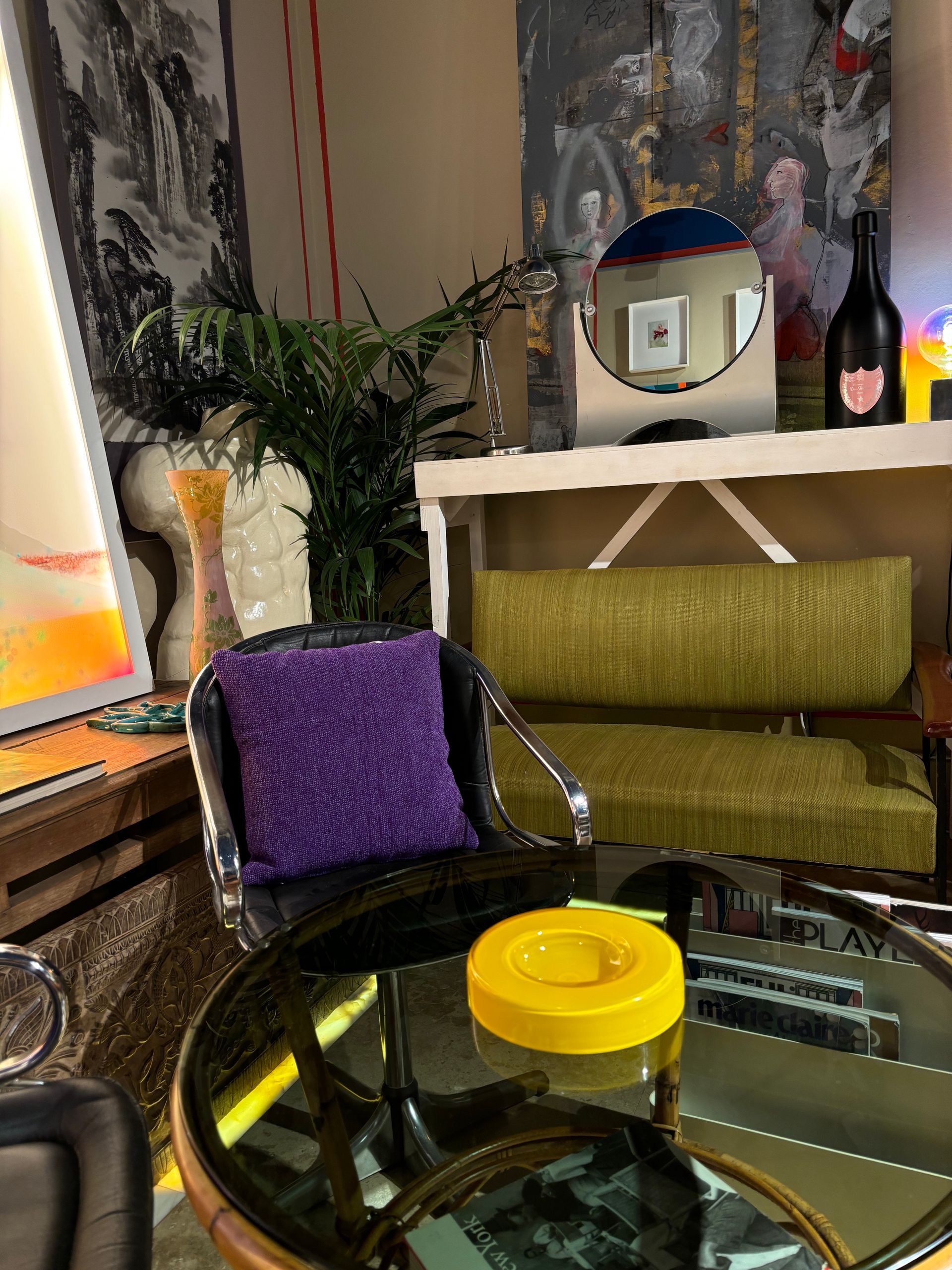 Nella stanza si trovano un cuscino viola su una sedia, un tavolino da caffè in vetro con un coperchio giallo e, sullo sfondo, un divano verde.