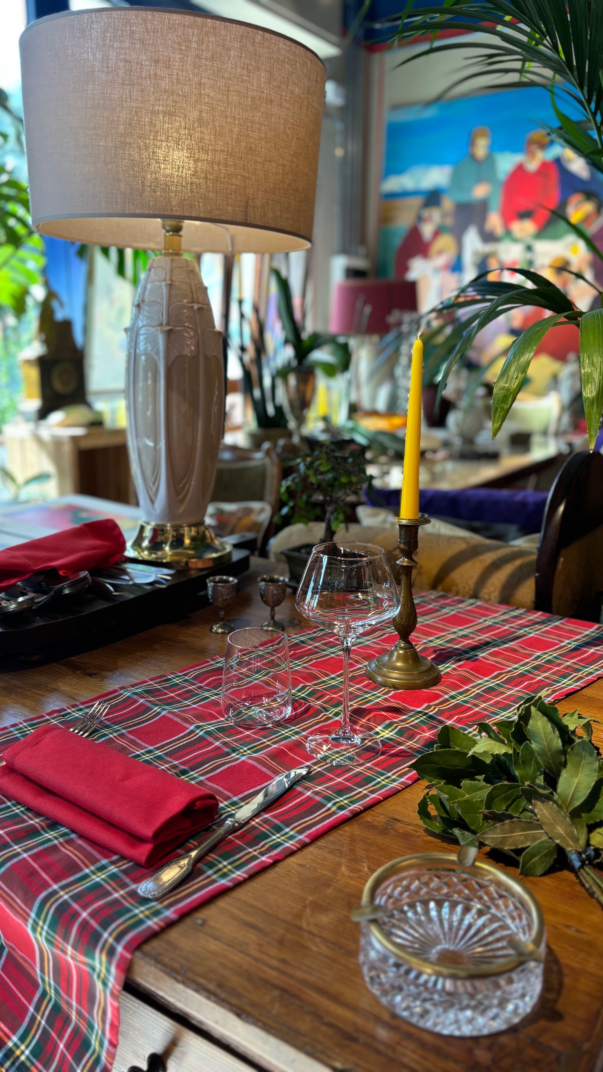 Una tavola apparecchiata per la cena con un runner a quadri, una lampada, una candela gialla, tovaglioli rossi, bicchieri e piante decorative.