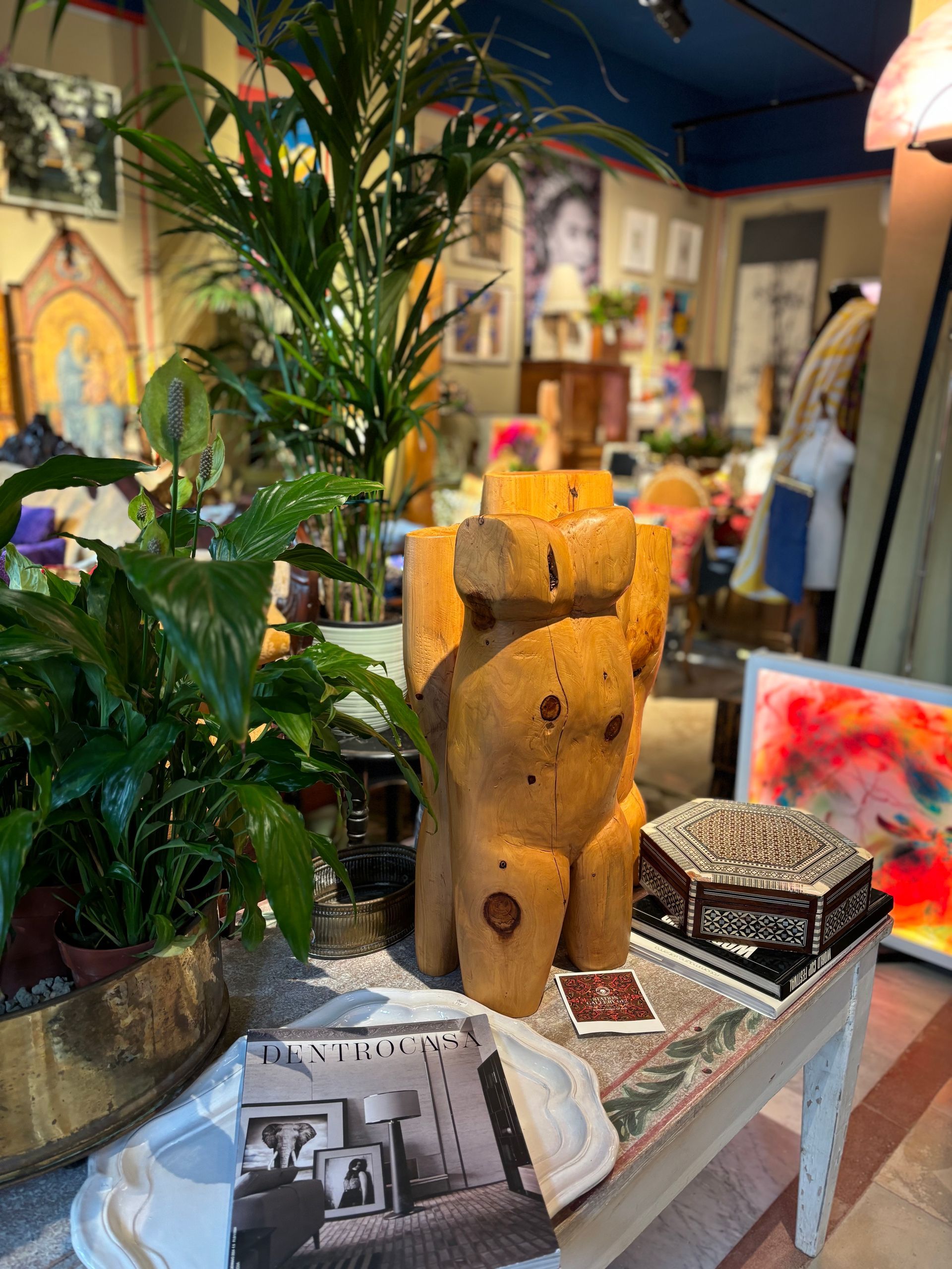 Una scultura in legno raffigurante un torso si erge su un tavolo accanto a una pianta verde e rigogliosa, una scatola decorata e un libretto stampato.