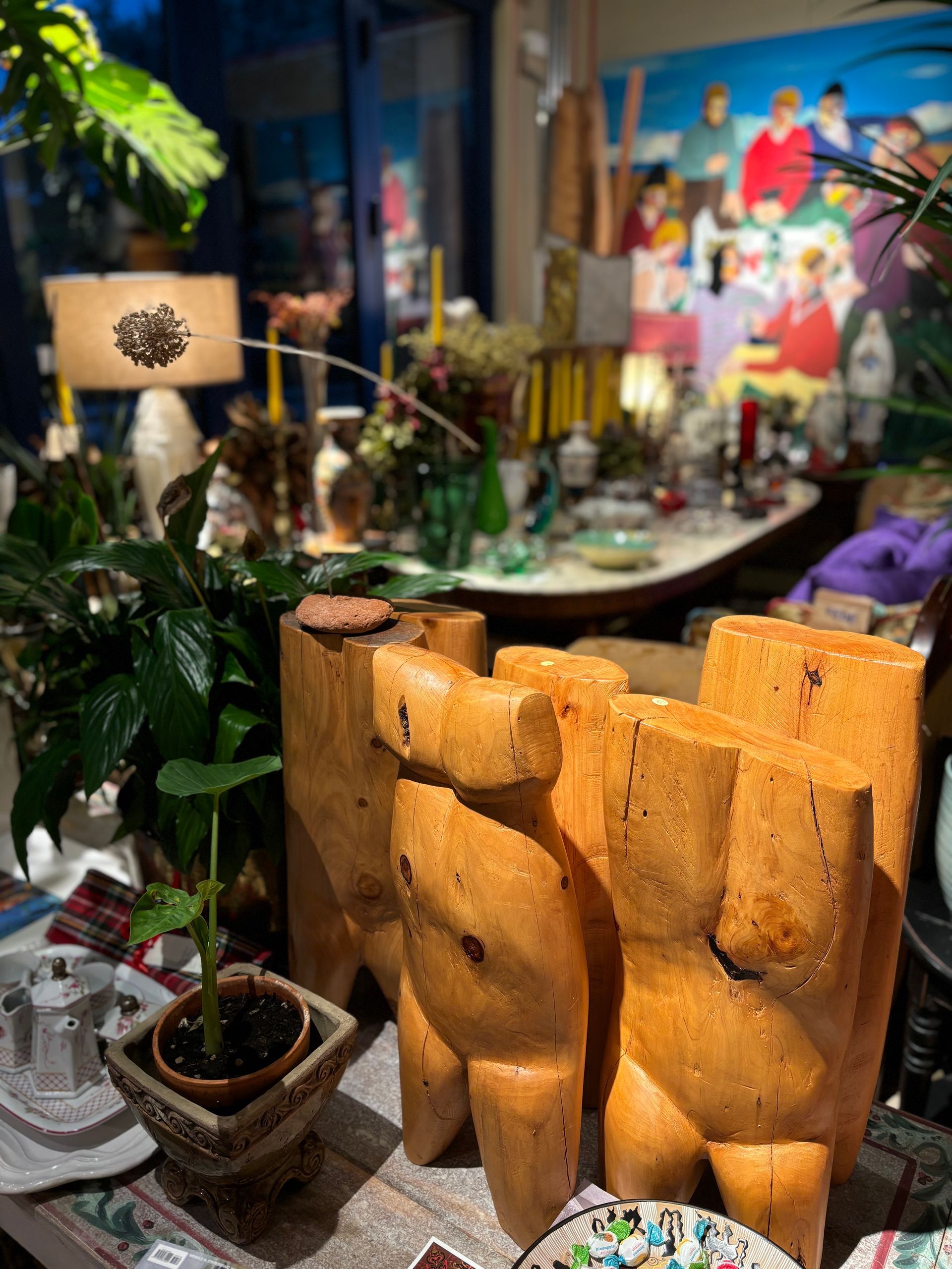 Due sculture stilizzate di busti in legno sono posizionate su un tavolo accanto a una pianta in vaso, con sullo sfondo un interno di negozio sfocato.