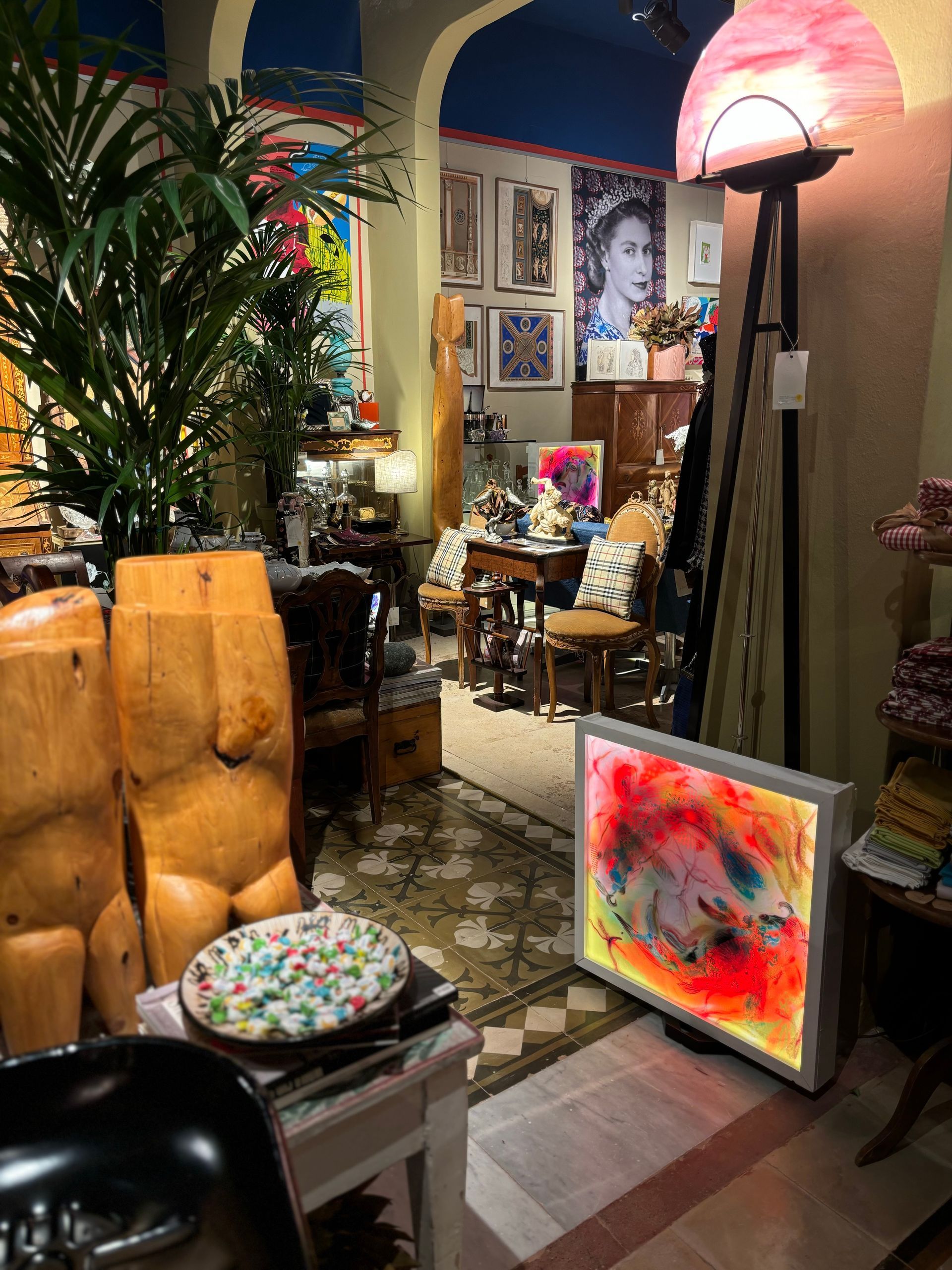 Un negozio interno disordinato, con sculture di busti in legno, una lampada con paralume rosa, una scatola luminosa colorata e stampe artistiche.