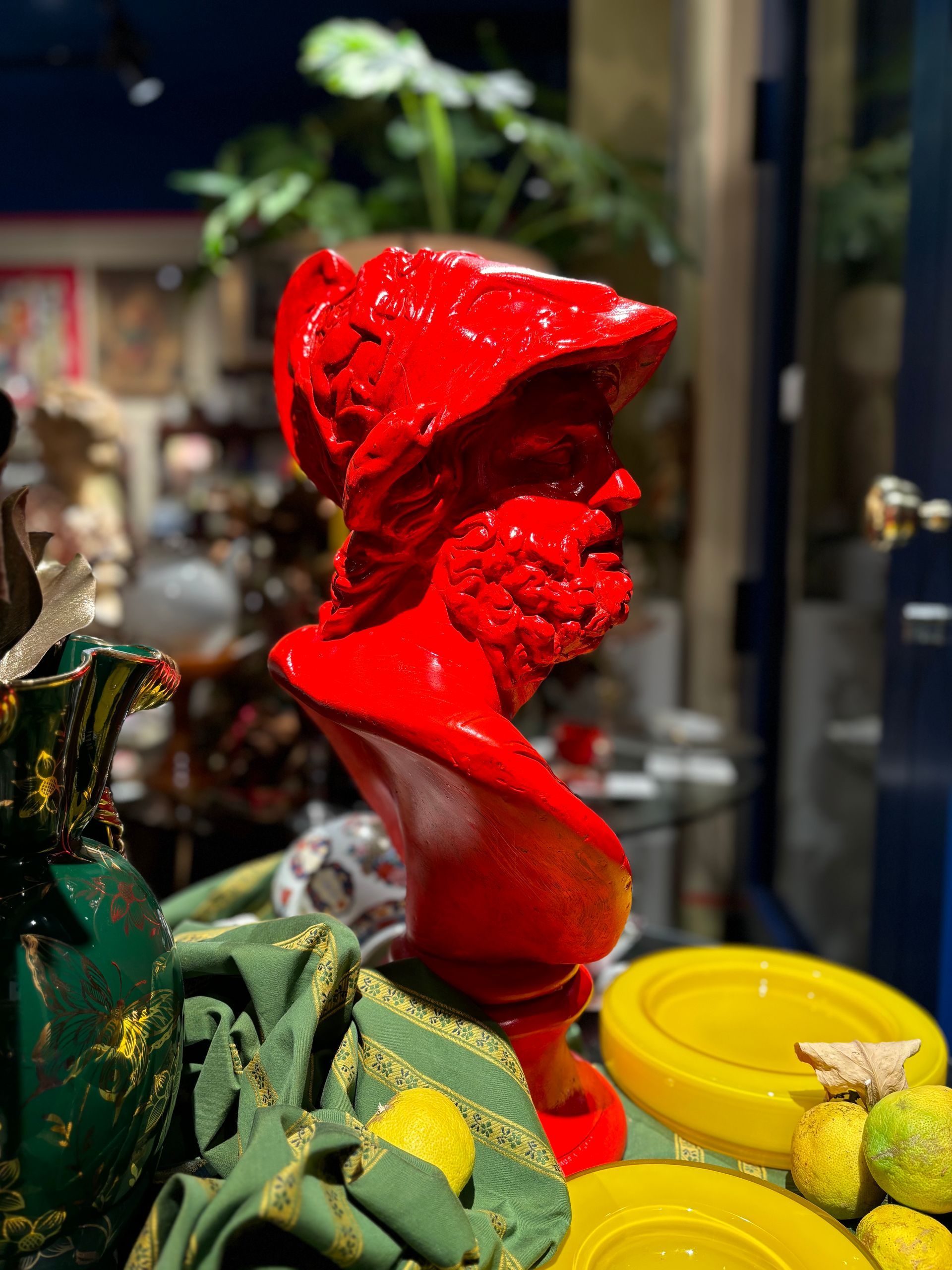 Un busto rosso acceso di un guerriero barbuto con elmo, posizionato tra oggetti decorativi in un negozio.