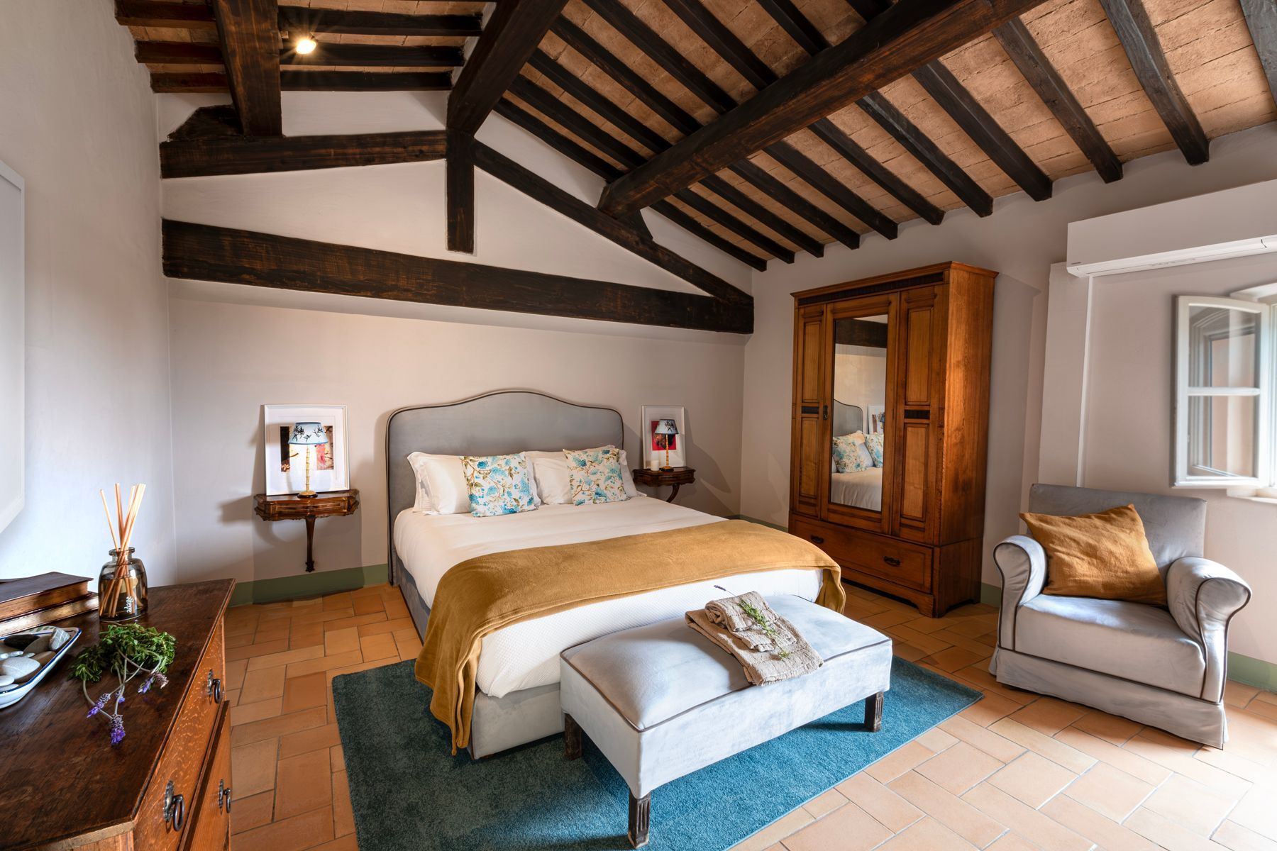 Una camera da letto rustica con travi in legno a vista, un letto matrimoniale, un armadio in legno, un tappeto blu e una poltrona grigia.