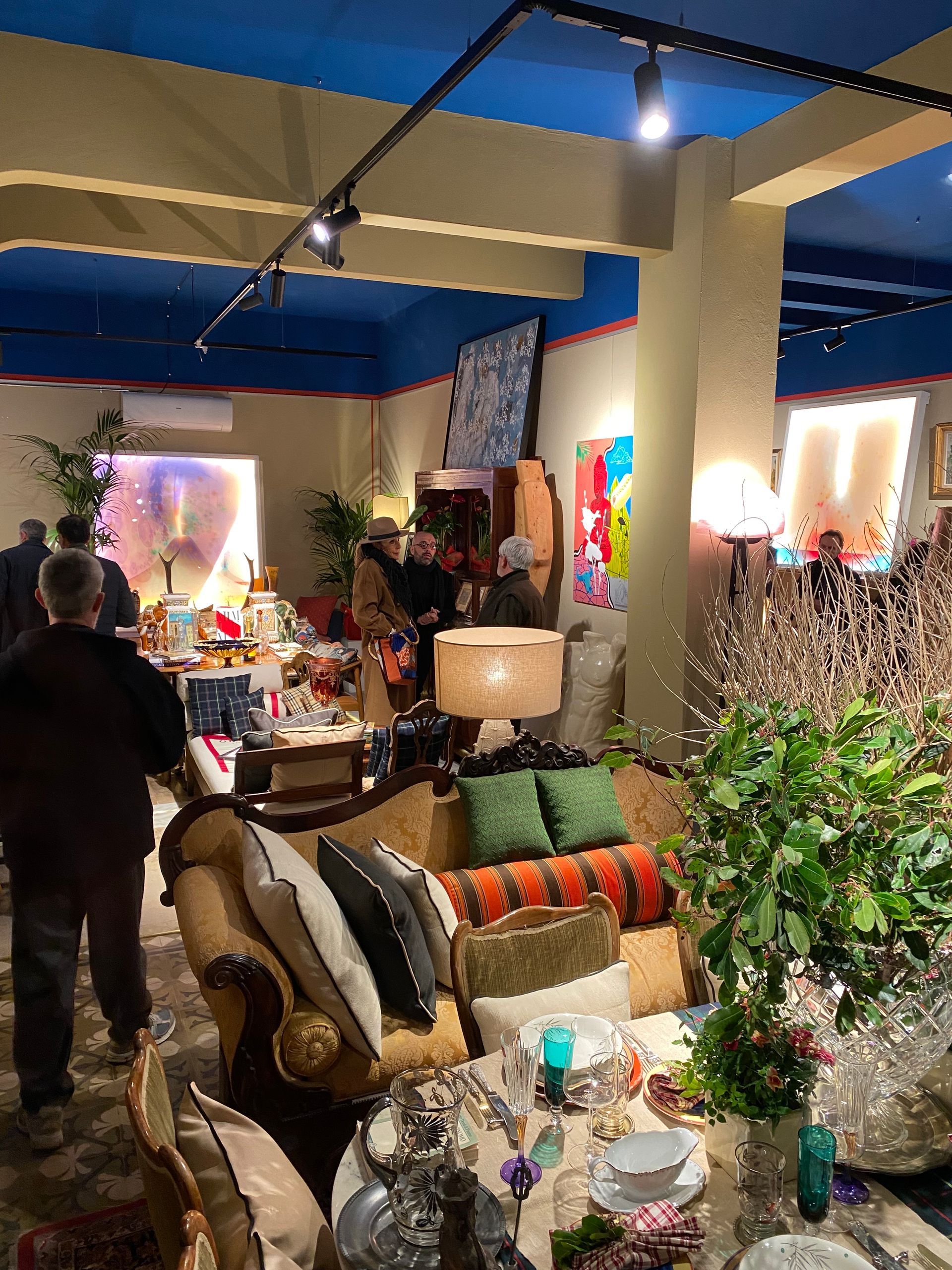 Un accogliente ritrovo al chiuso in una stanza illuminata da una luce calda e ricca di opere d'arte, con un divano, un tavolo e persone che chiacchierano.