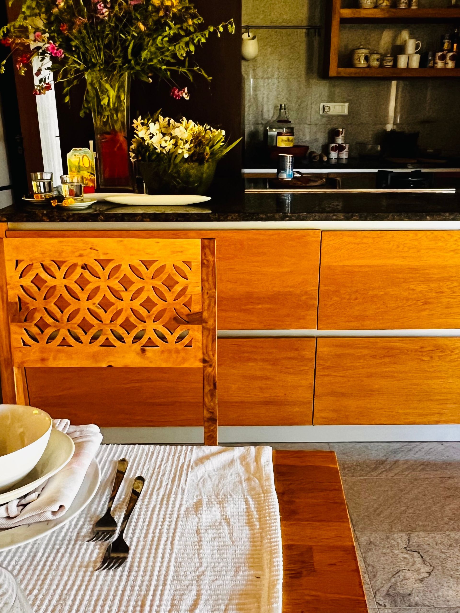 Un tavolo da pranzo con le posate è posizionato di fronte a un bancone da cucina in legno dalle tonalità calde, decorato con un motivo a griglia.