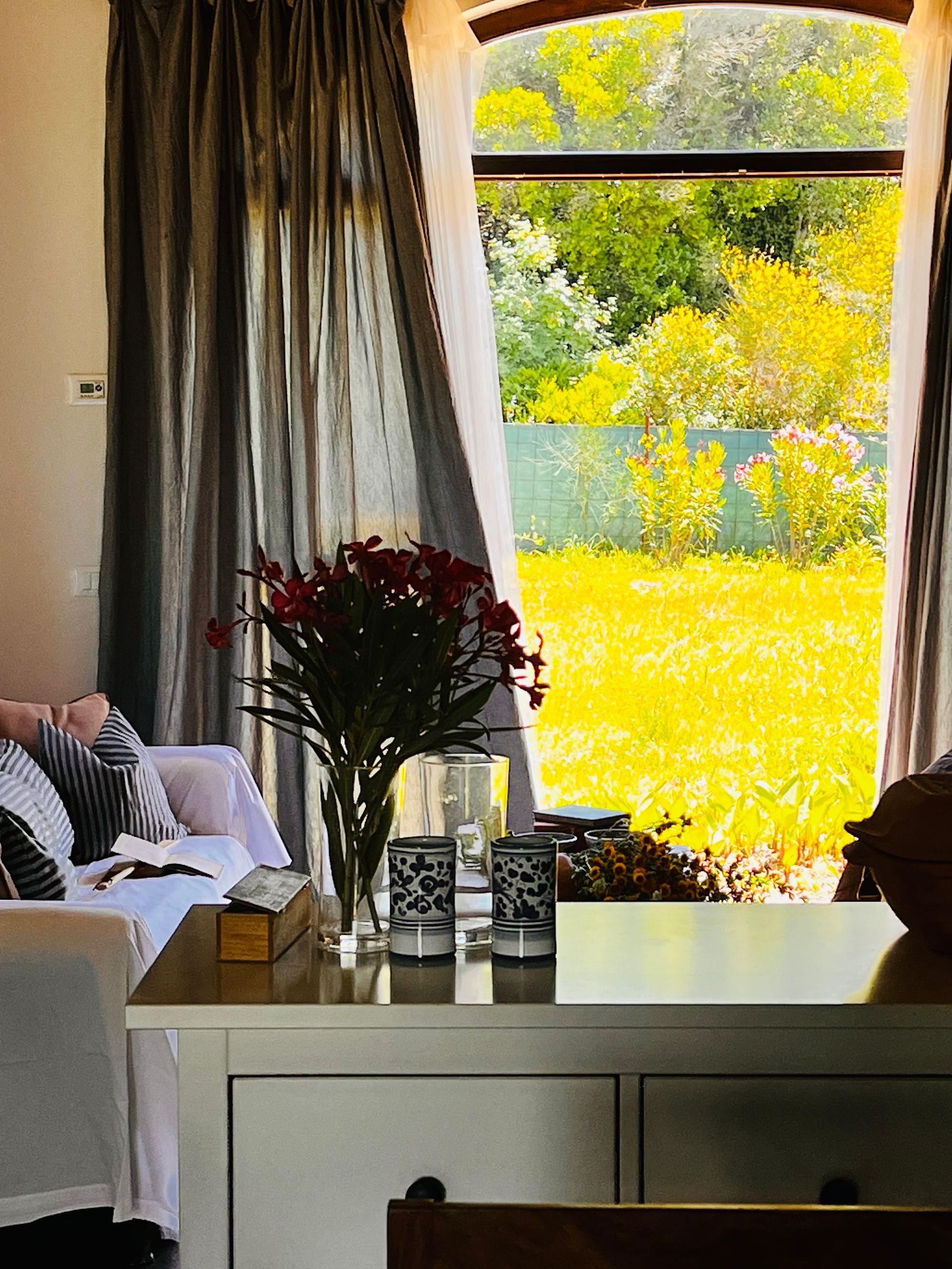 Un vaso di fiori rossi è appoggiato su un tavolo davanti a una finestra che si affaccia su un giardino soleggiato e giallo.