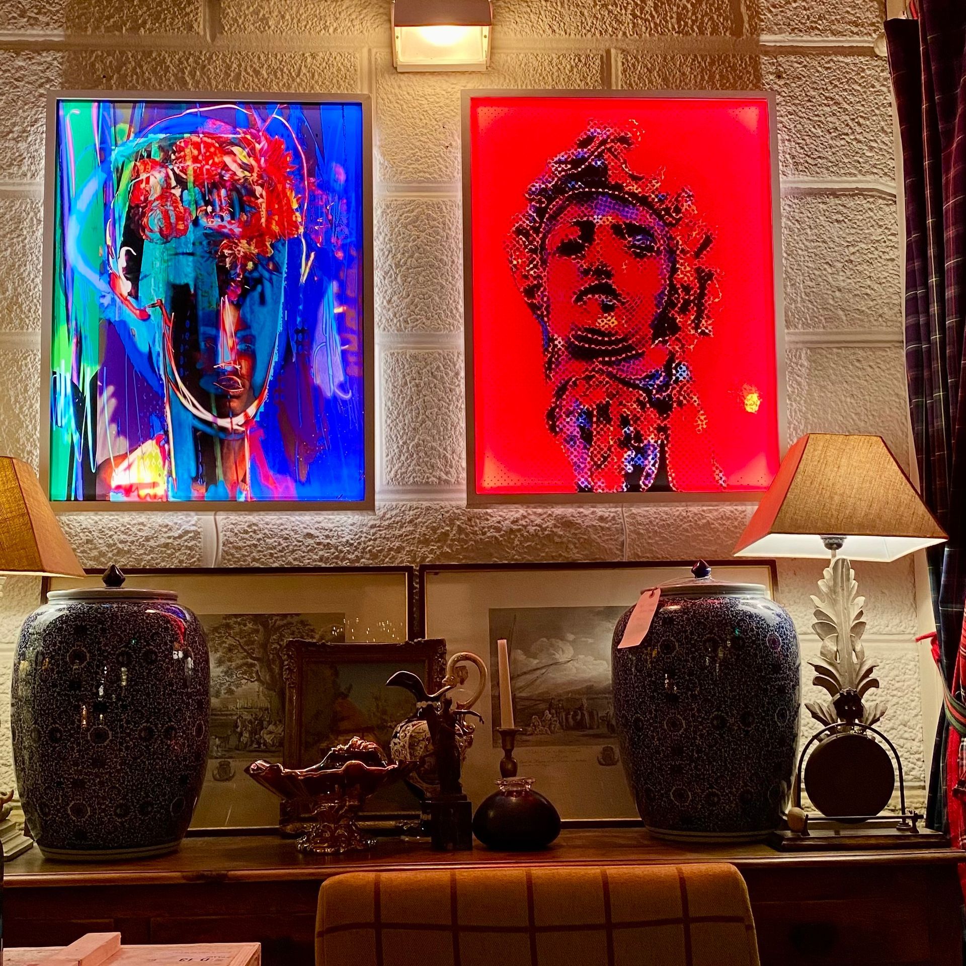 Sopra una consolle in legno, ornata da due grandi urne in ceramica blu scuro e una lampada, sono appesi due ritratti moderni e dai colori vivaci.