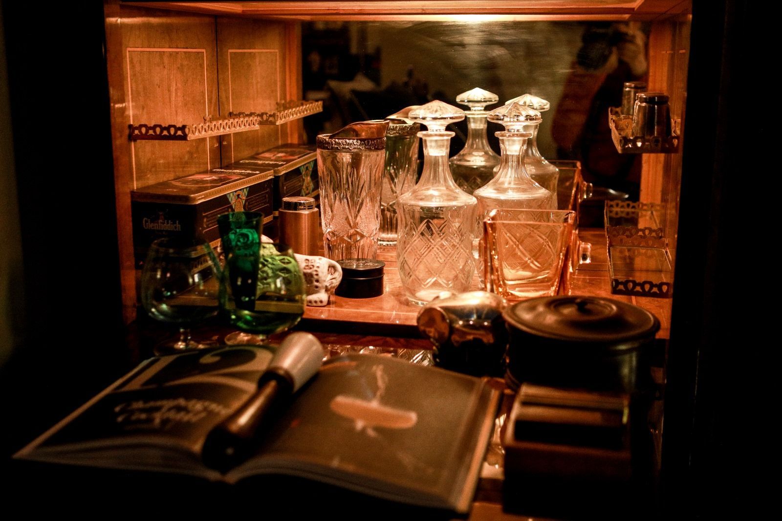 Uno scaffale da bar con caraffe e bicchieri di cristallo, con un libro di cocktail aperto e un pestello in primo piano.