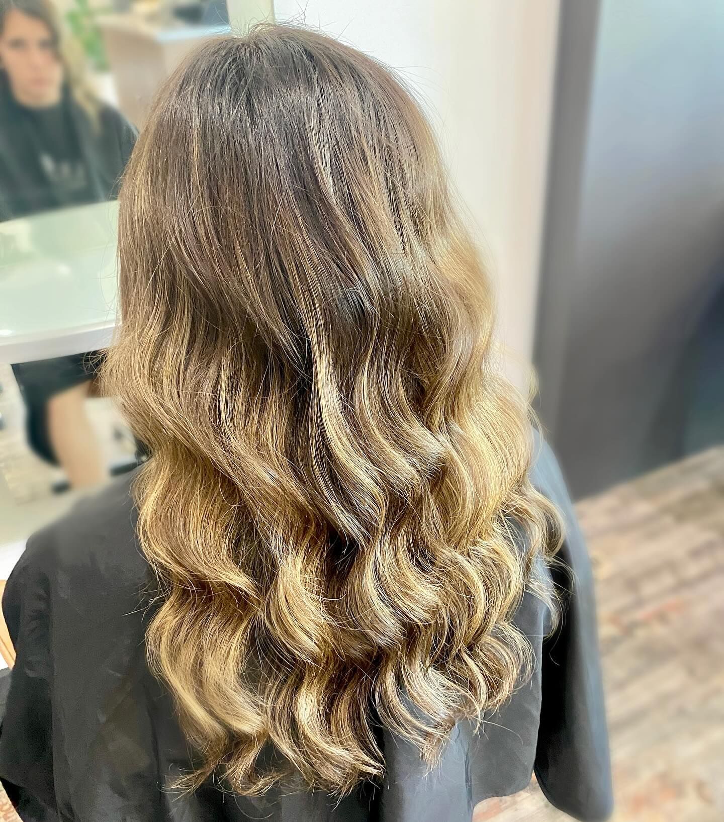 A woman with long wavy brown hair is sitting in a salon chair — Carlee Hair Design In Anula, NT