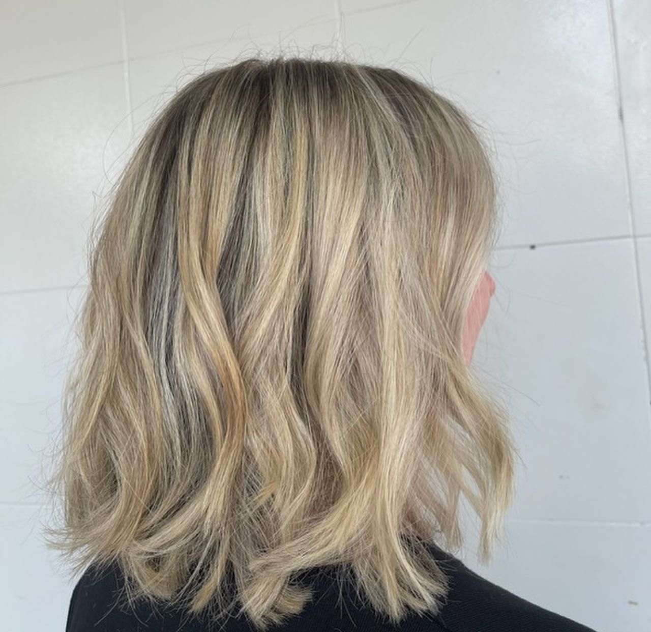 A woman with short curly blonde hair is standing in front of a white brick wall — Carlee Hair Design In Anula, NT