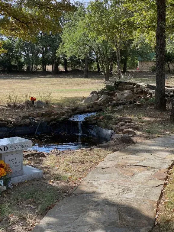 A stone path leads to a small garden pond with a waterfall, located near a memorial bench and trees in a grassy field.