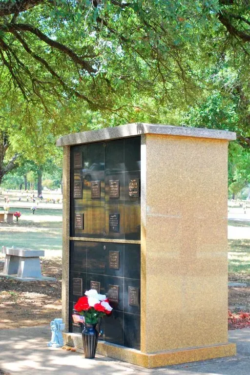 A tan stone columbarium with black niches for cremated remains in a park-like cemetery with a vase of flowers in front.