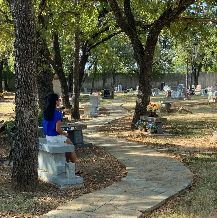 A person in a blue shirt sits on a stone bench along a winding stone path in a sunny, wooded cemetery.