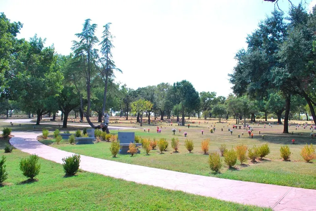 A landscaped cemetery park with trees, grassy plots, grave markers, and a winding paved walkway under a clear sky.