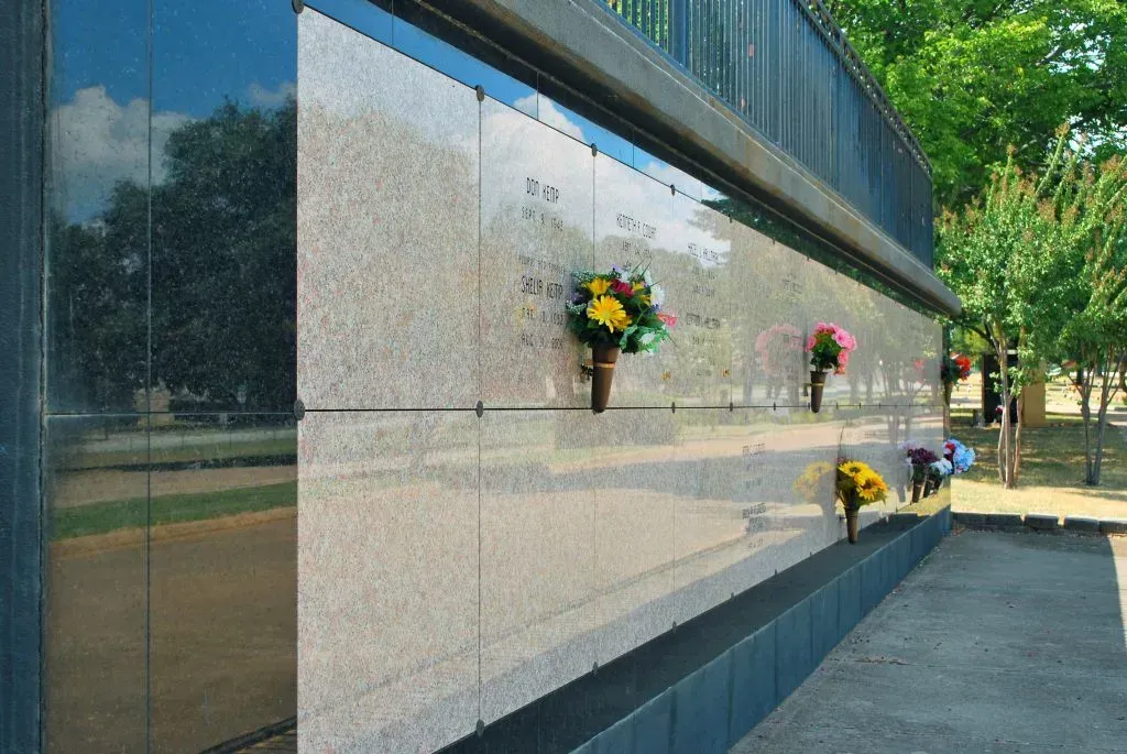 A columbarium wall made of light polished stone, featuring multiple niches with small floral bouquets, in an outdoor park.