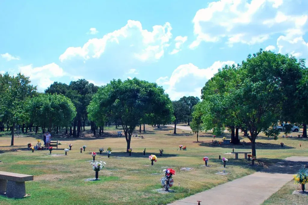 A cemetery on a sunny day featuring flat grave markers with floral arrangements, scattered trees, and a concrete path.