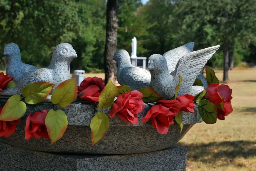 Stone doves perched on a round, gray stone basin adorned with red silk roses in a cemetery.