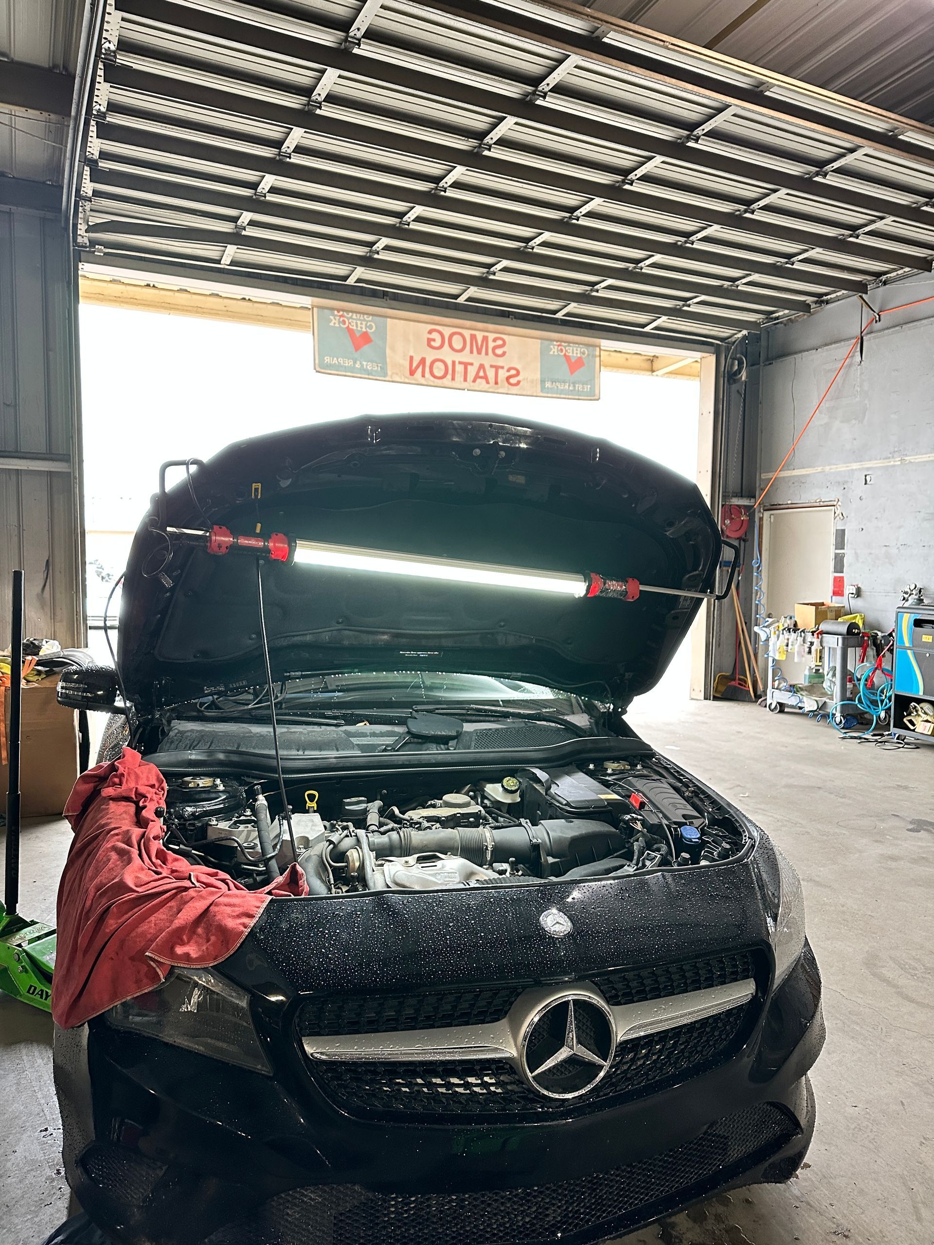 Black car with open hood inside a repair shop. Red shop towels and light bar are visible.