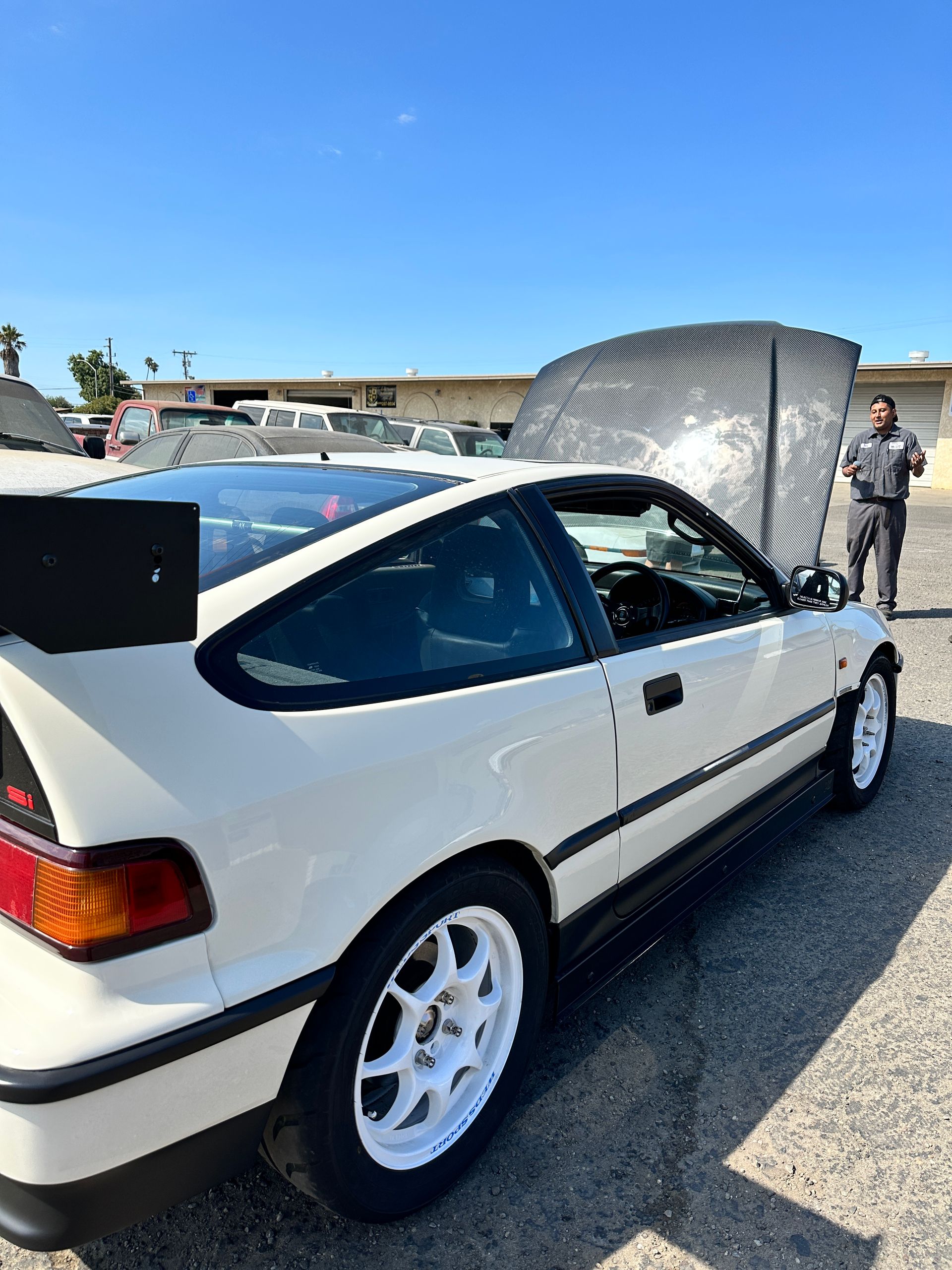 White Honda CRX with open hood, black spoiler, and a person standing next to it under a blue sky.