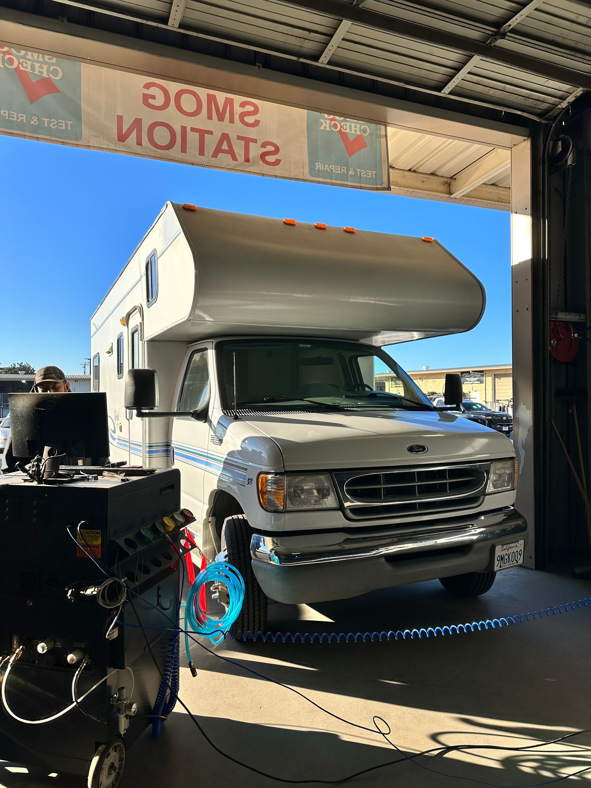 RV inside a smog check station. A blue fan blows air toward the front tires. 