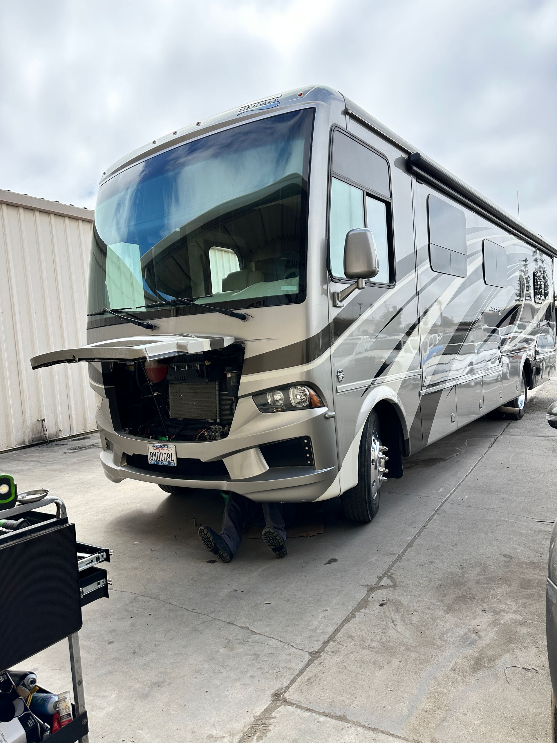 Beige and gray RV parked outside a building, front bumper removed, visible engine components.