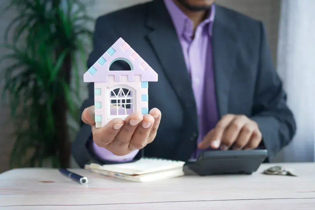A ballpoint pen, a notebook, a calculator, and a man wearing corporate attire in the background holding a puzzled house tool.