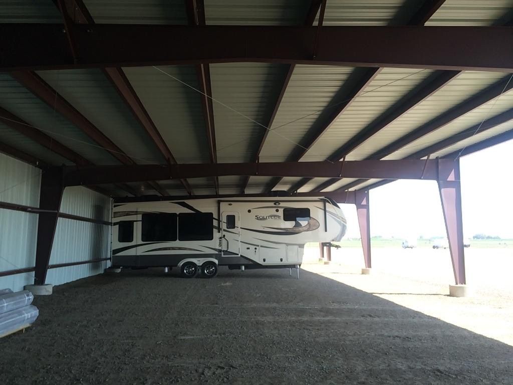 White RV parked under a metal-roofed open shed with gravel ground and bright daylight outside