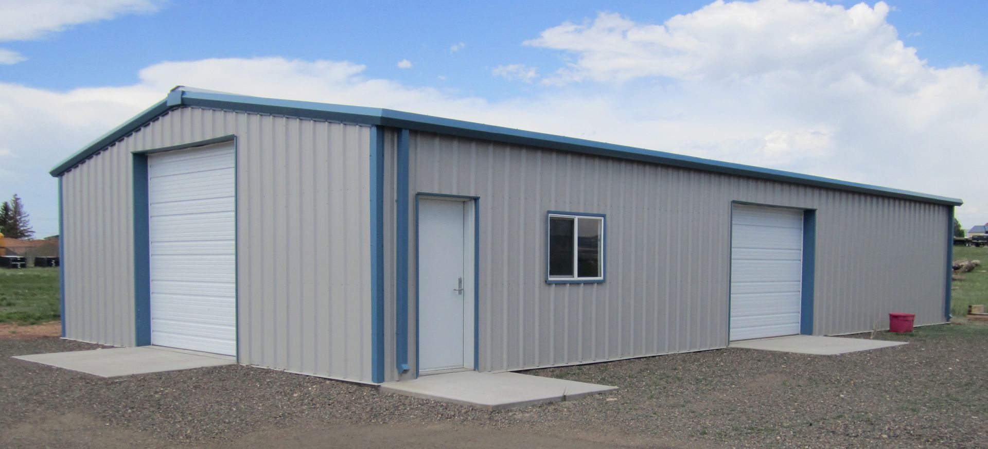 Gray metal warehouse with blue trim and roll-up doors on a gravel lot