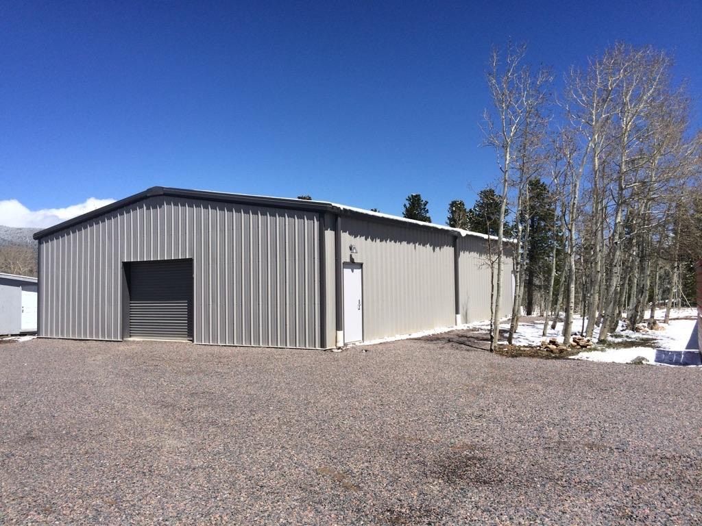 Gray metal warehouse with large garage door on a gravel lot beside snowy trees under a clear blue sky