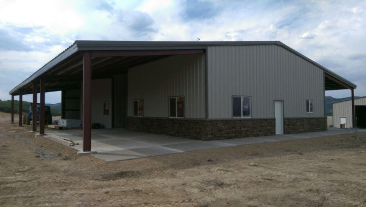 Large gray metal warehouse with a covered porch and stone base on a dirt lot under a cloudy sky