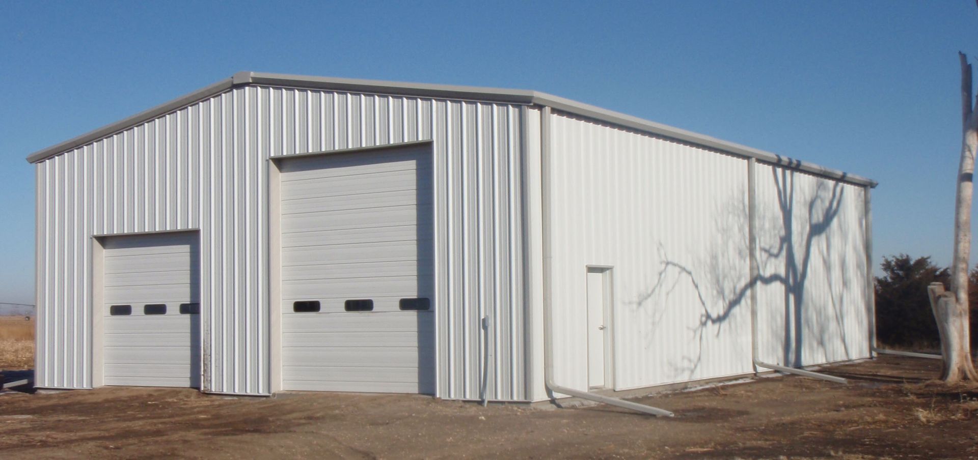 White metal warehouse with two garage doors on a dirt lot under a clear blue sky
