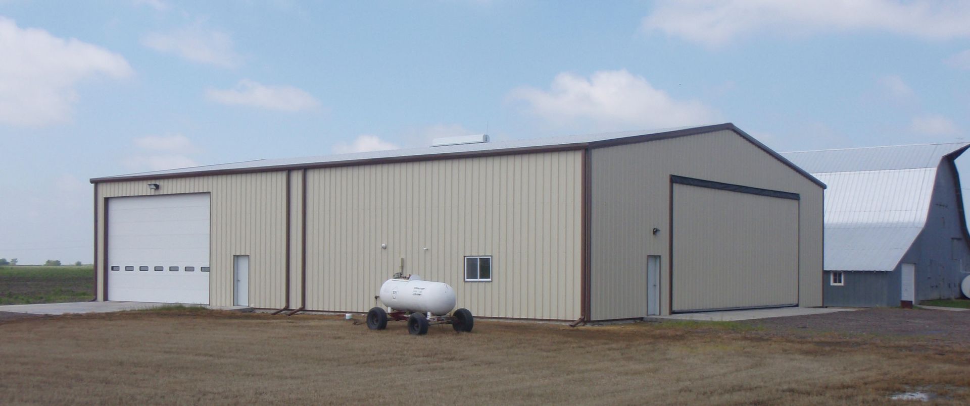 Large beige metal warehouse with garage doors in a rural field under a cloudy sky.