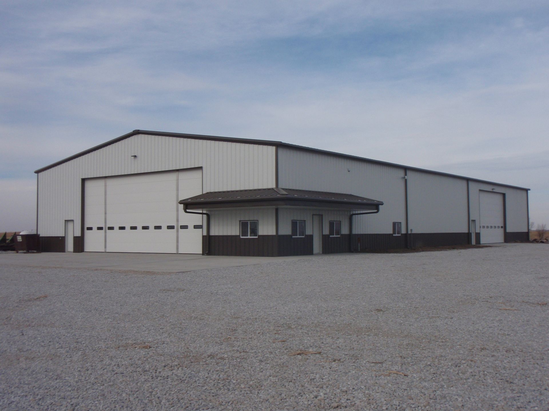 Large white metal warehouse with a small covered entrance on a gravel lot under a cloudy sky