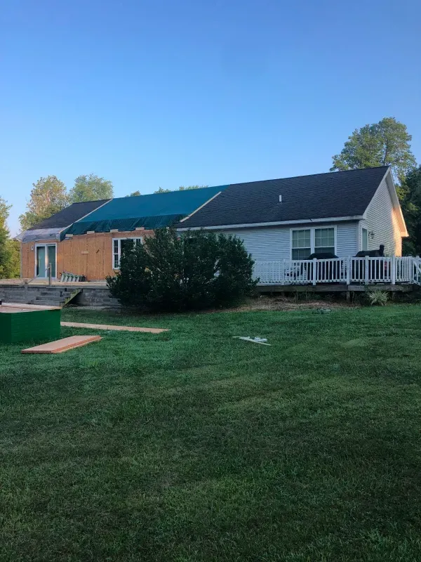Suburban house with a partially tarp-covered roof, white porch, and grassy yard under a blue sky