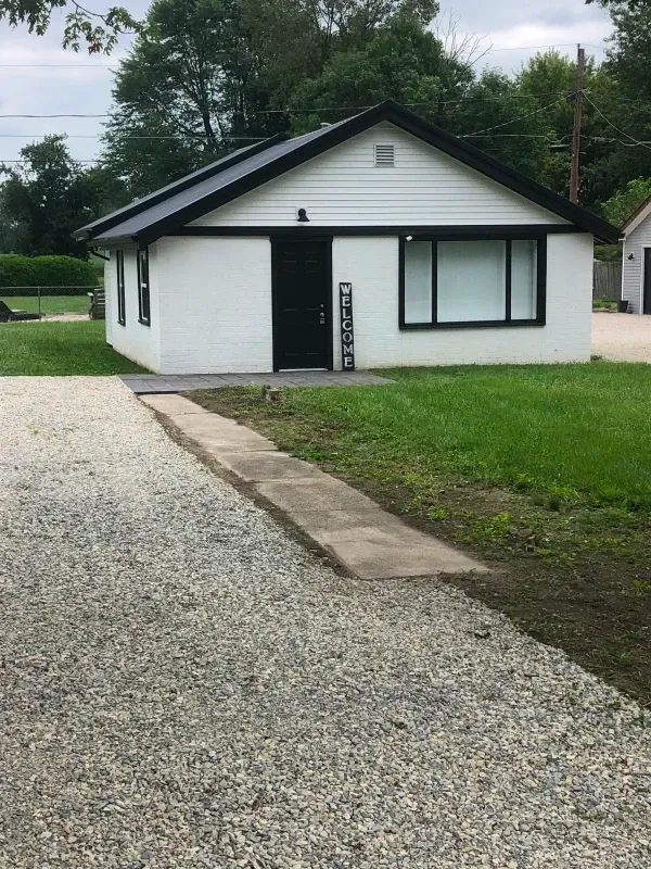 Small white house with black trim beside a gravel driveway and lawn under cloudy skies.