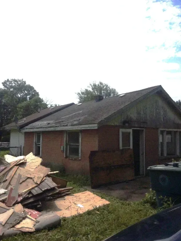 Damaged brick house with roof shingles torn off and debris piled in the front yard