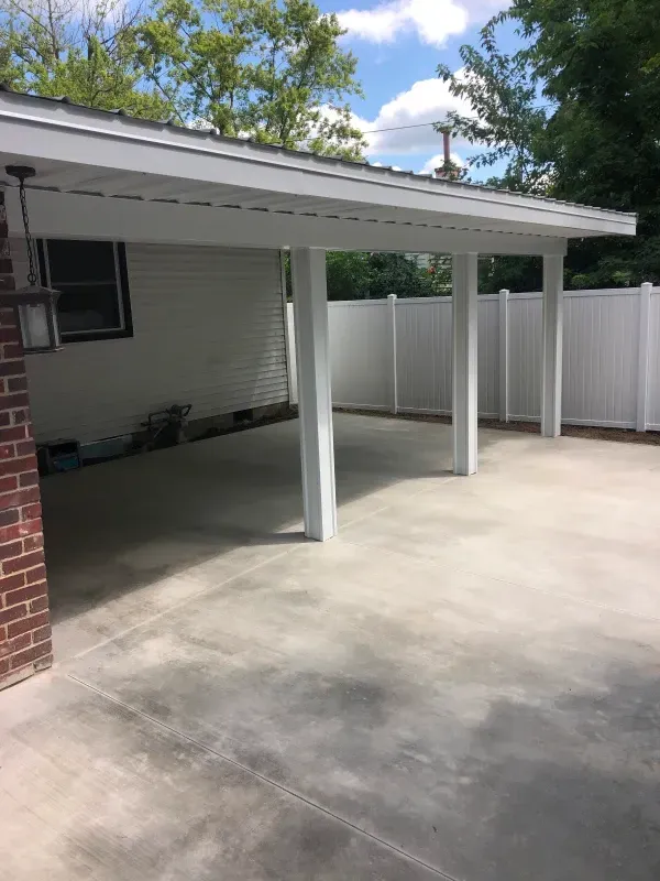 Covered concrete patio with white posts and fence beside a house under a partly cloudy sky